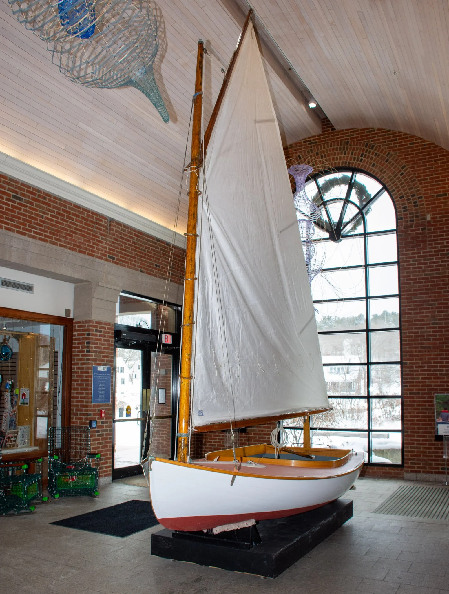 An indoor display of a small sailboat with white hull, orange trim, and beige sail, situated on a black platform inside a building with brick walls and large arched window.