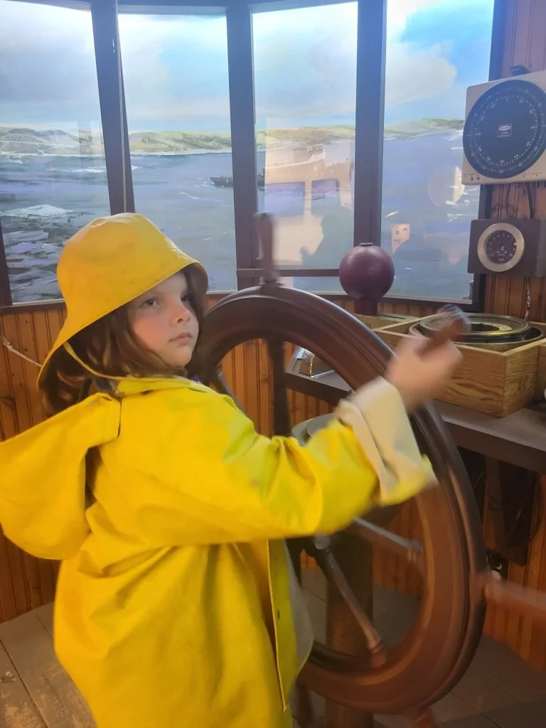 A young girl wearing a yellow raincoat and rain hat steering a ship's wheel on a boat exhibit with a window view of water and distant land.