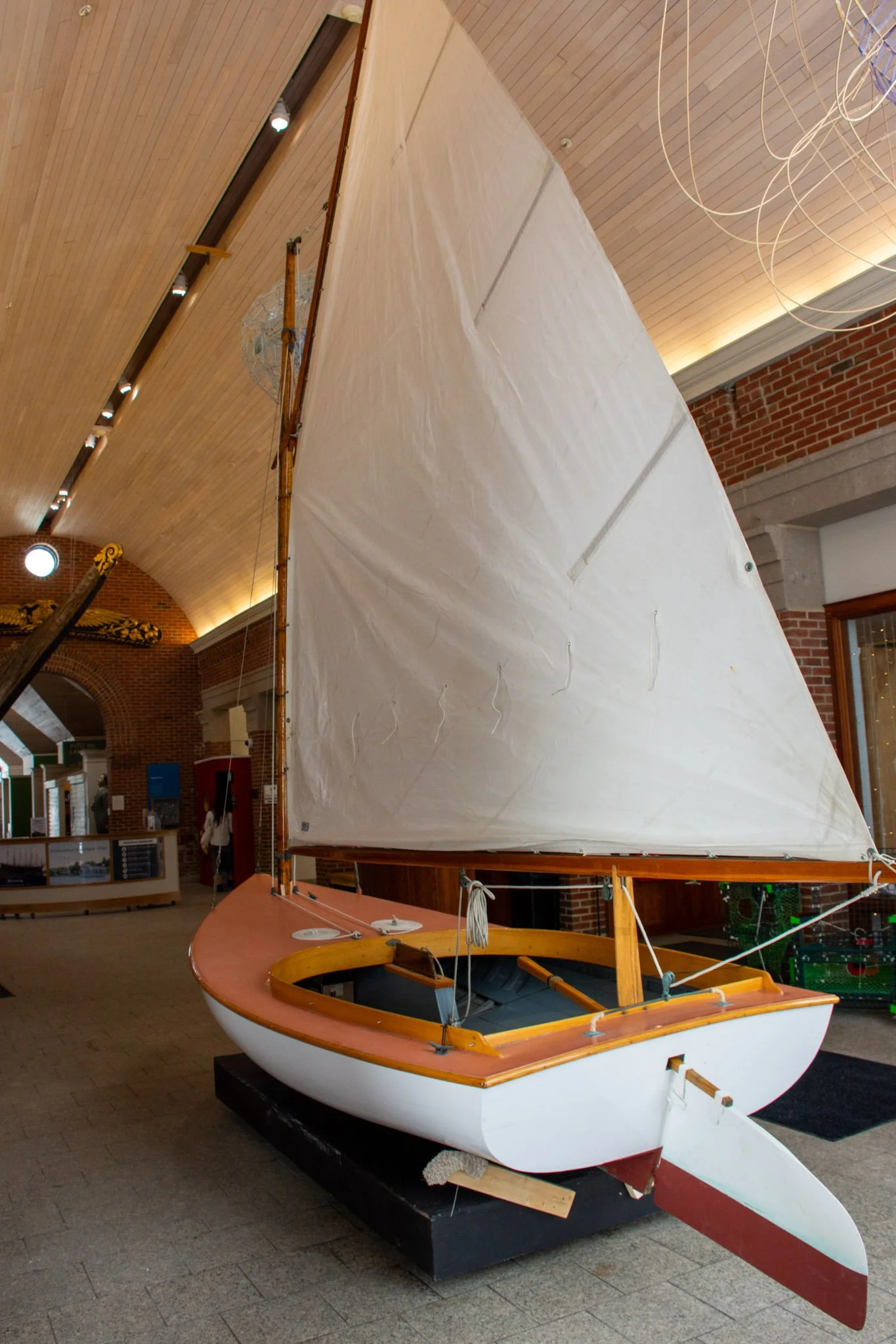 A sailboat with a white hull and wooden trim displayed indoors, with a large white sail and a wooden tiller, in a room with brick walls and a wooden ceiling.