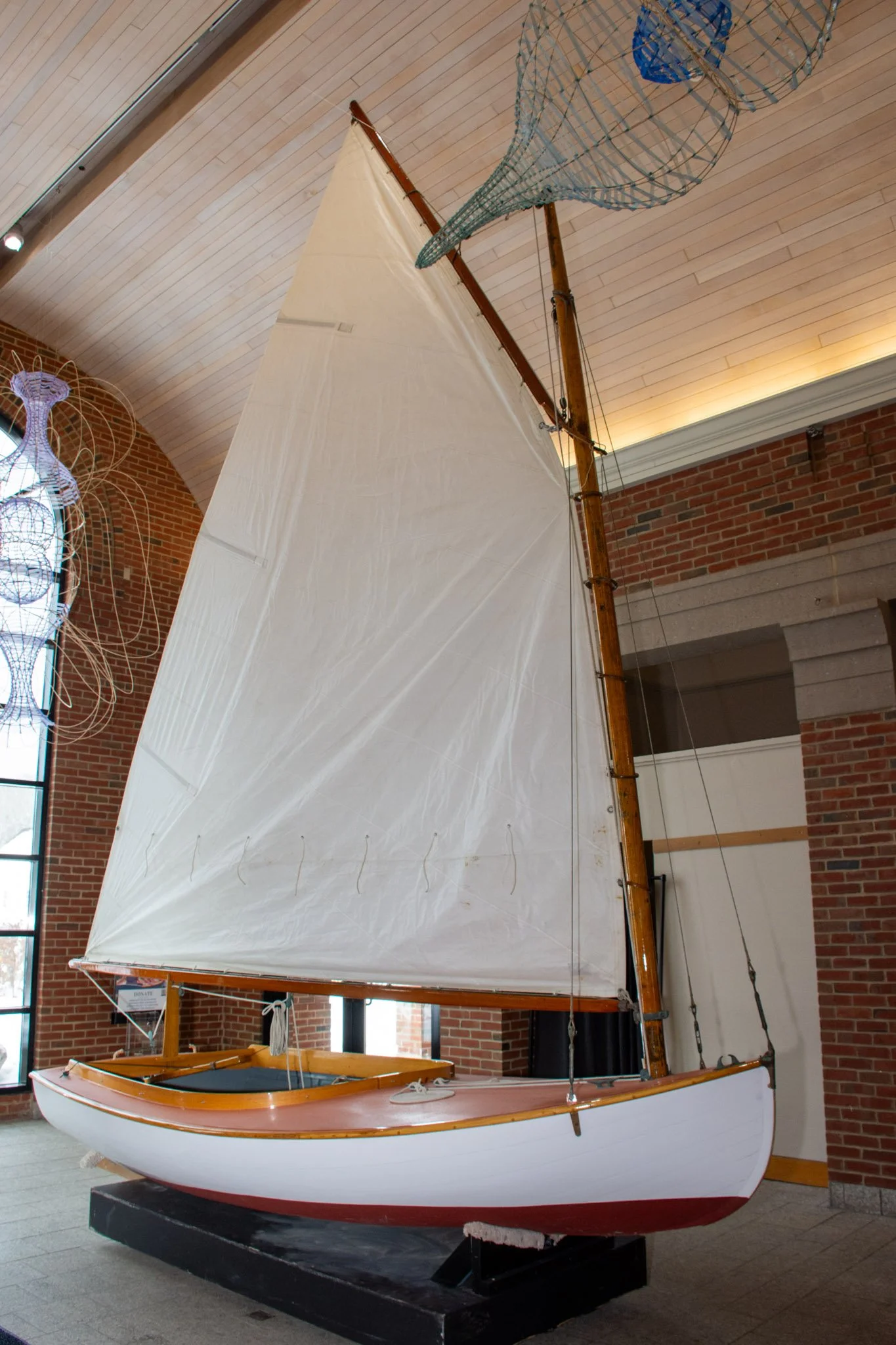 A model sailboat displayed indoors with a wooden hull, white deck, and tall white sail, situated on a black stand against brick and beige wall background.