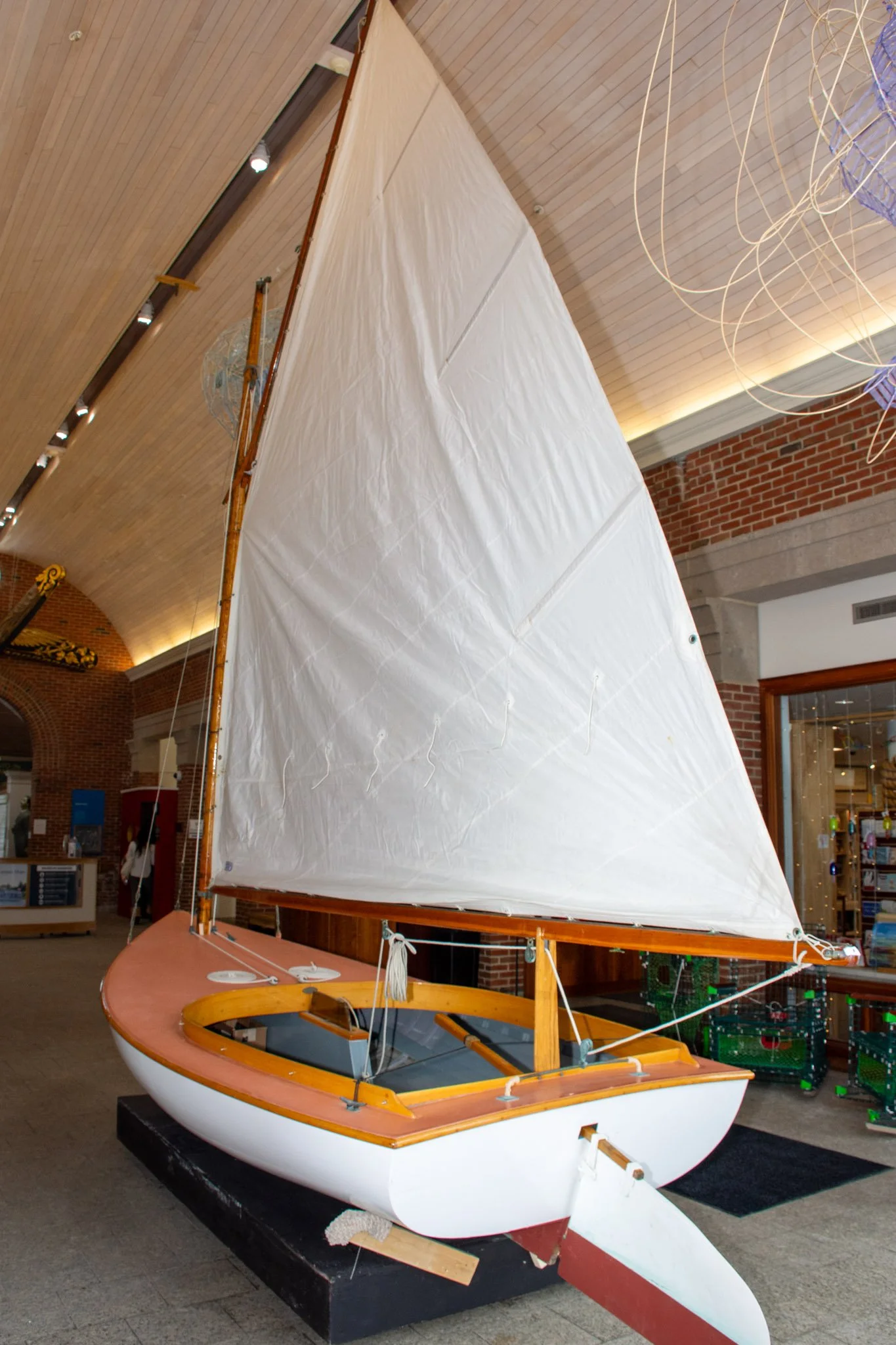 A small sailboat with a brown wooden deck and a white hull, featuring a large white sail, displayed indoors on a black platform. The background shows a brick wall, windows, and decorative lighting.