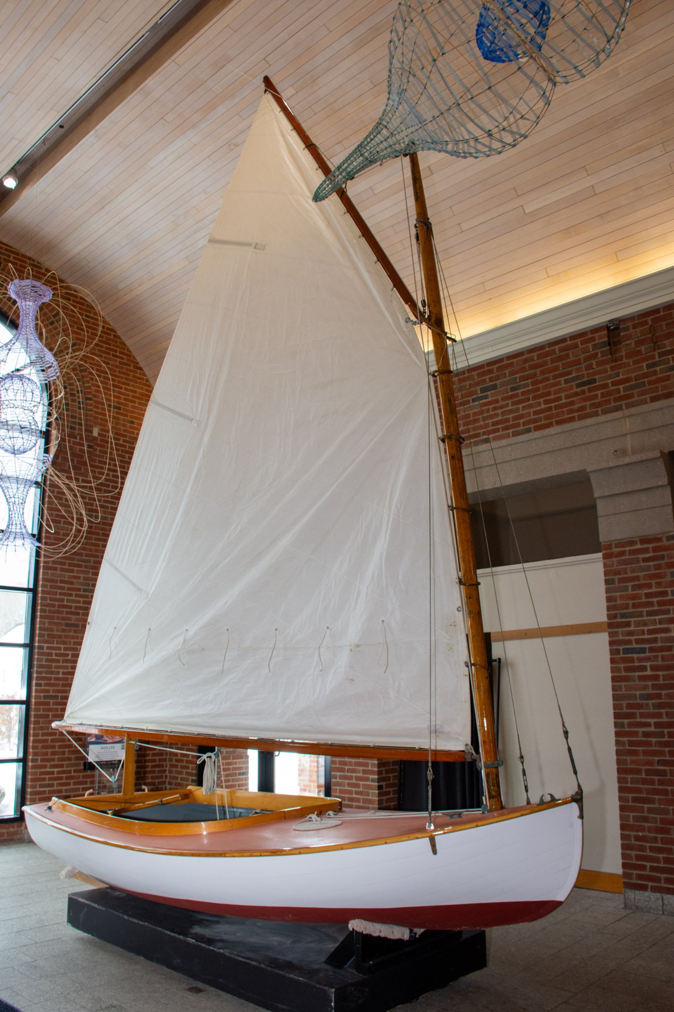 A sailboat with a white hull, wooden trim, and a large white sail is displayed indoors on a black stand. The boat has a wooden mast and a green fins and a blue-green fishing net is hanging from the ceiling. The room has brick walls and a wooden ceiling.