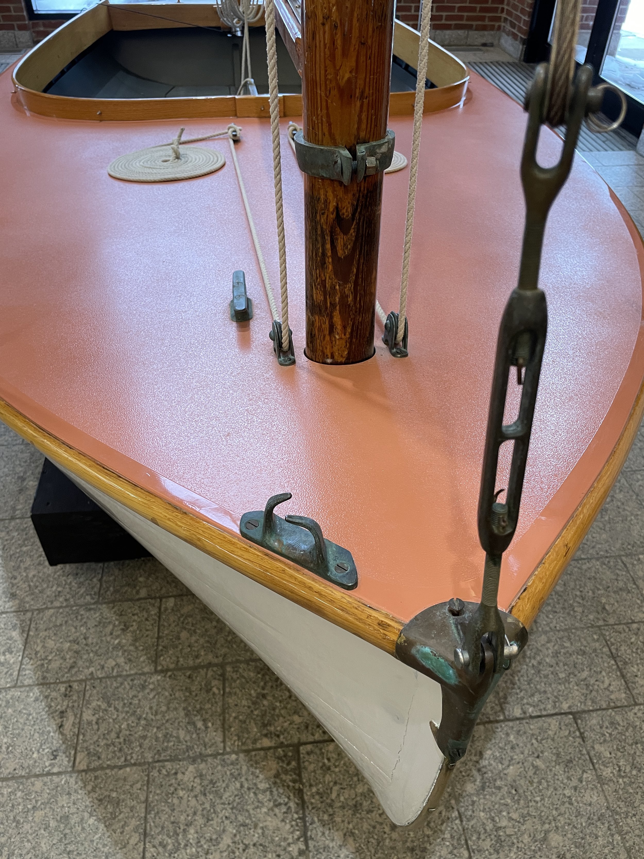 Image of a small wooden boat with a pink textured surface, metal fastenings, ropes, and a wooden mast, placed indoors on a tiled floor.