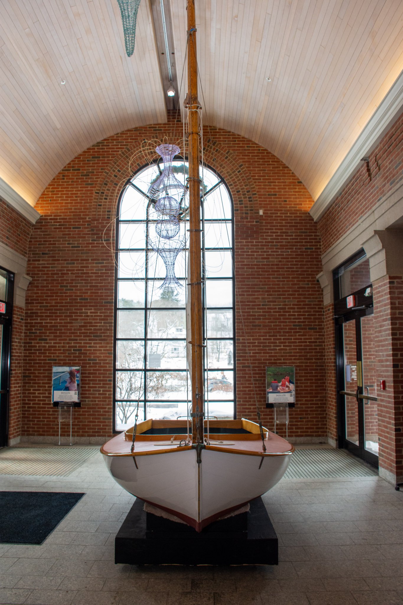 A boat with a wooden mast and rigging displayed indoors against brick walls and a large window, with informational boards nearby.