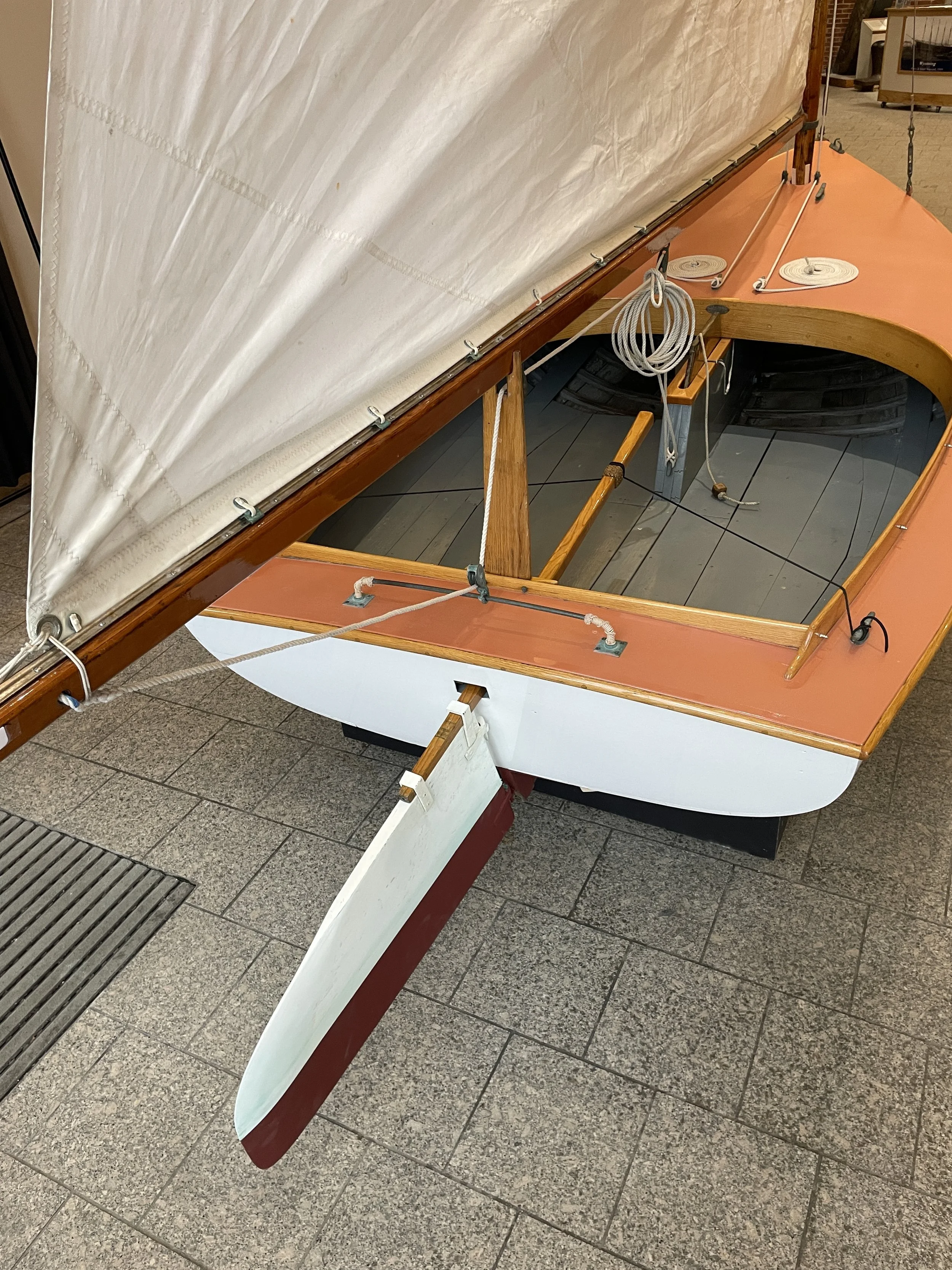 A wooden boat on display indoors, with its sail partially raised and ropes and rigging visible inside and on the side.