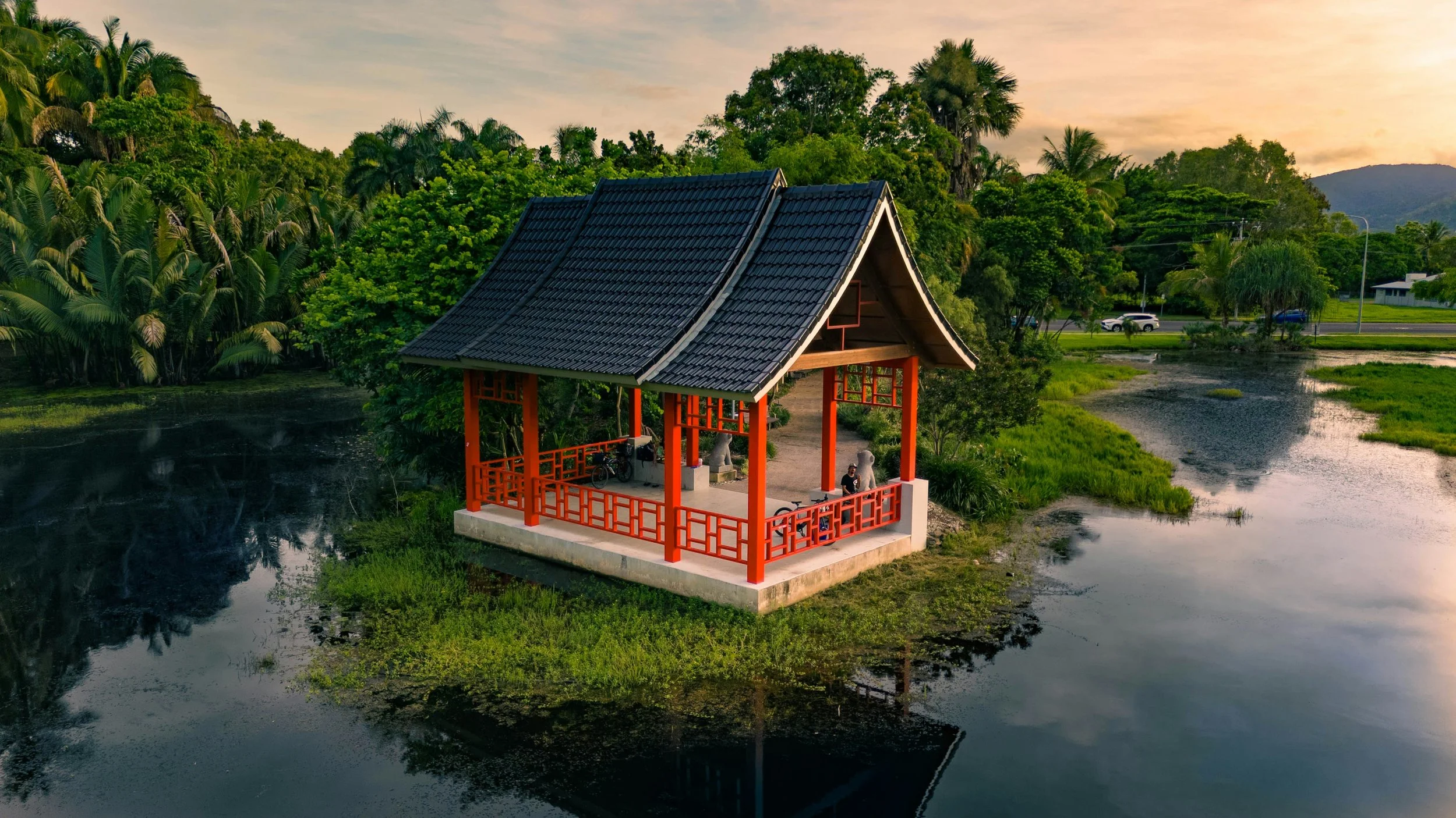 Cairns Chinese Friendship Garden red Pavilion standing atop the water with a sunset and lush gardens in the background.