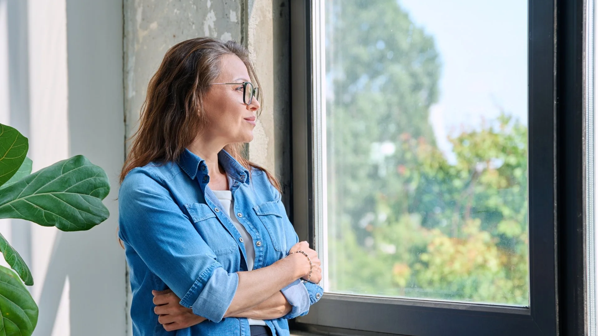 A woman with glasses and long brown hair standing indoors by a window, looking outside with her arms crossed, wearing a blue denim shirt, next to a large green plant.