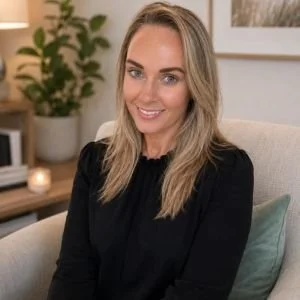 Portrait of a woman with blonde hair smiling, sitting on a beige couch in a cozy room with a plant and framed pictures on the wall.