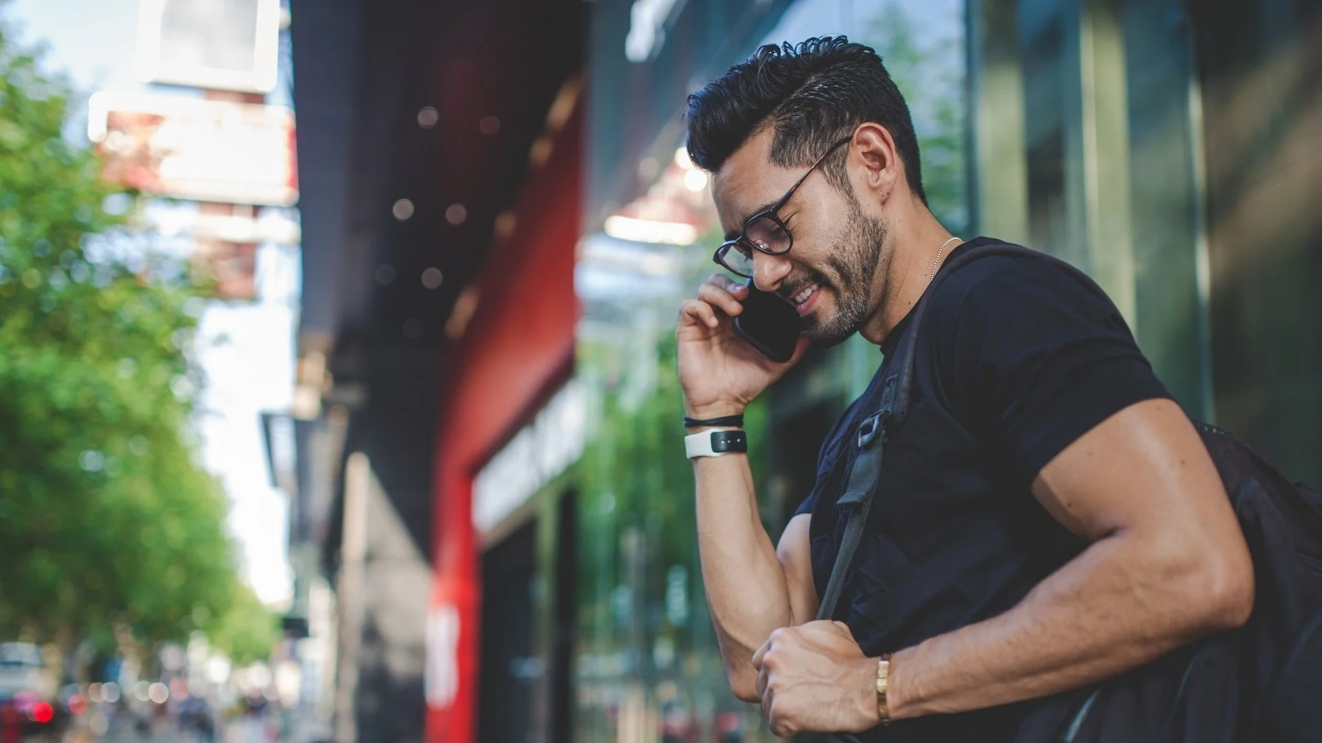 A man with glasses, dark hair, and a beard stands outside talking on his cell phone, smiling, with a backpack on, near a city street with trees and buildings in the background.
