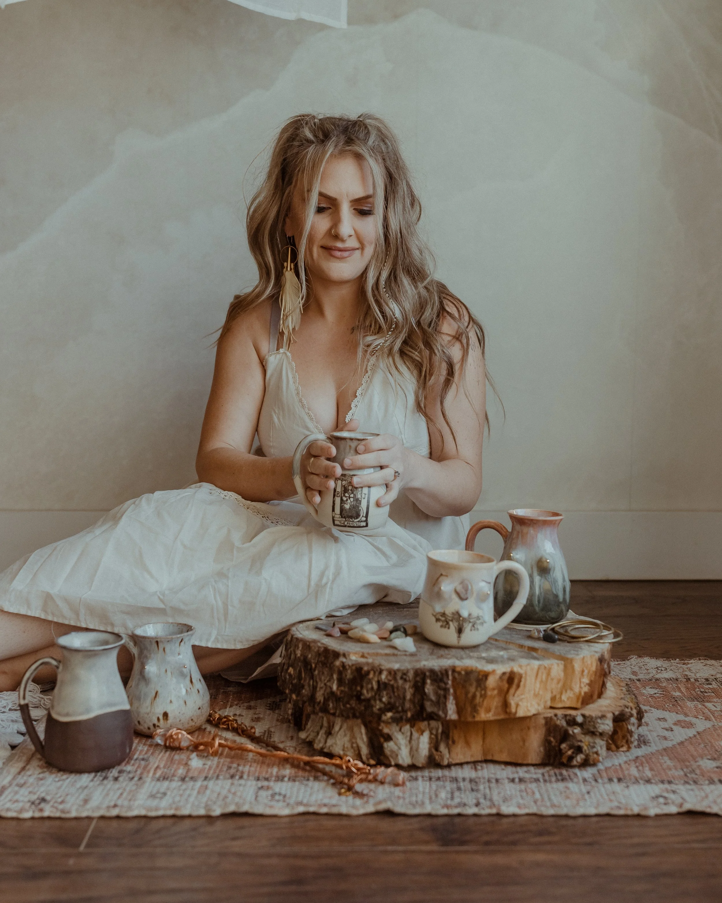 A woman sitting cross-legged on a patterned rug, holding a mug, surrounded by various rustic ceramic mugs and decorative stones on a wooden slab, with a neutral-colored wall in the background.