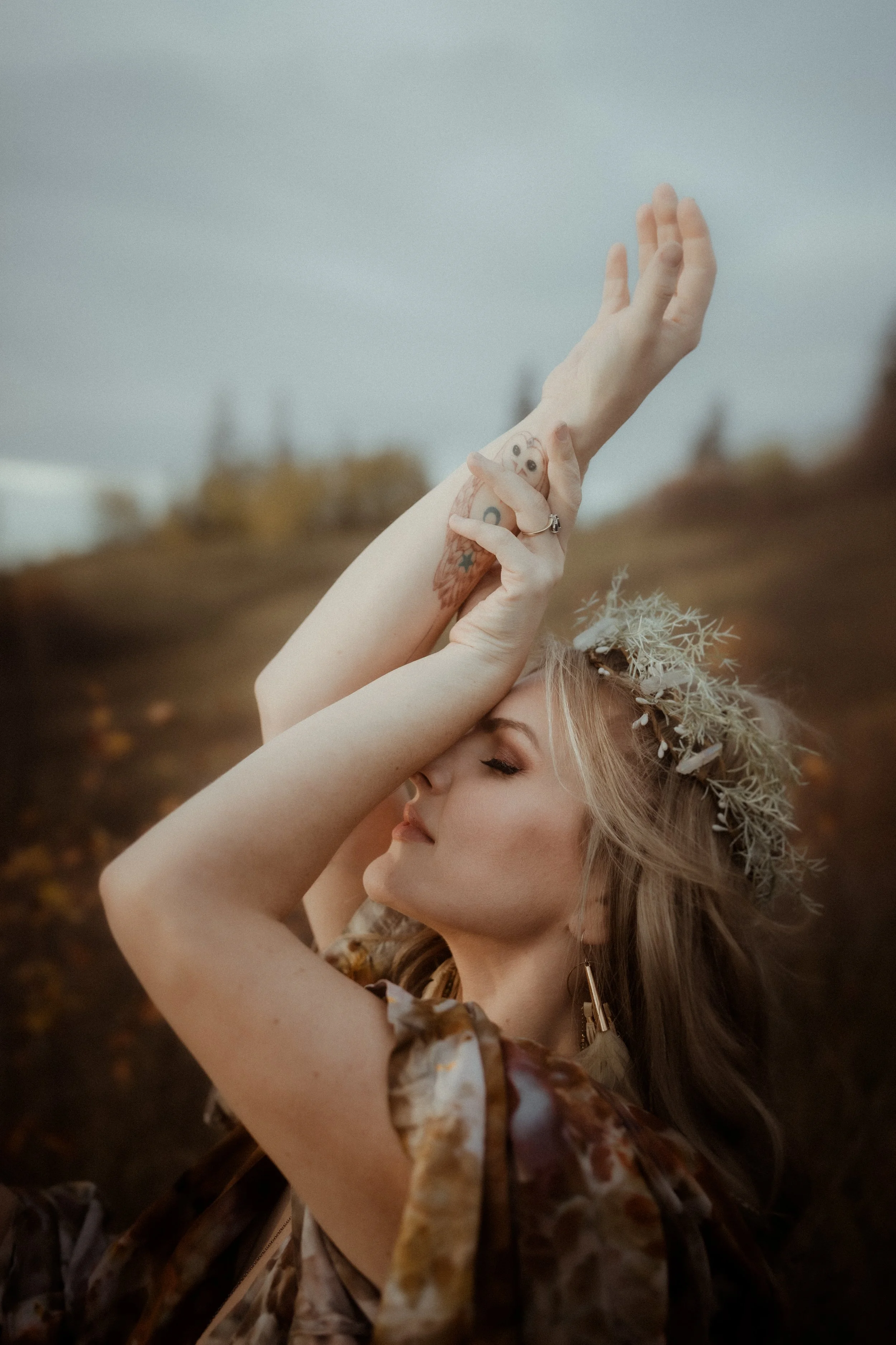 A woman with closed eyes and a serene expression, wearing a floral dress and a crown made of white foliage, is posing outdoors with her arms raised and crossed above her head, against a blurred natural landscape background.