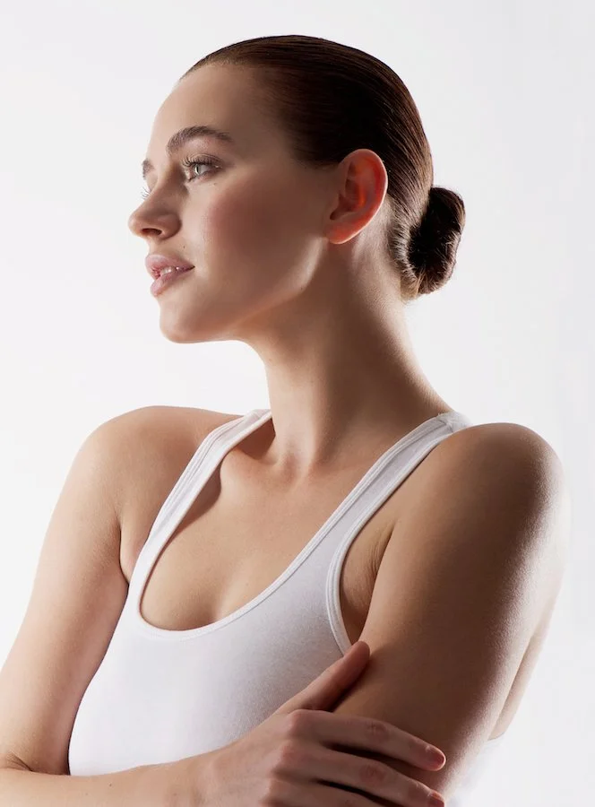 Profile of a young woman with brown hair tied in a bun, wearing a white tank top, looking to her left with arms crossed.