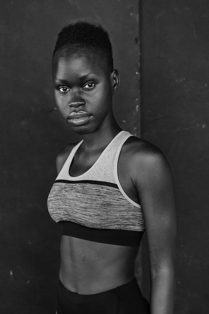Black and white photo of a young woman with a short haircut, wearing a sports bra, standing against a dark background.