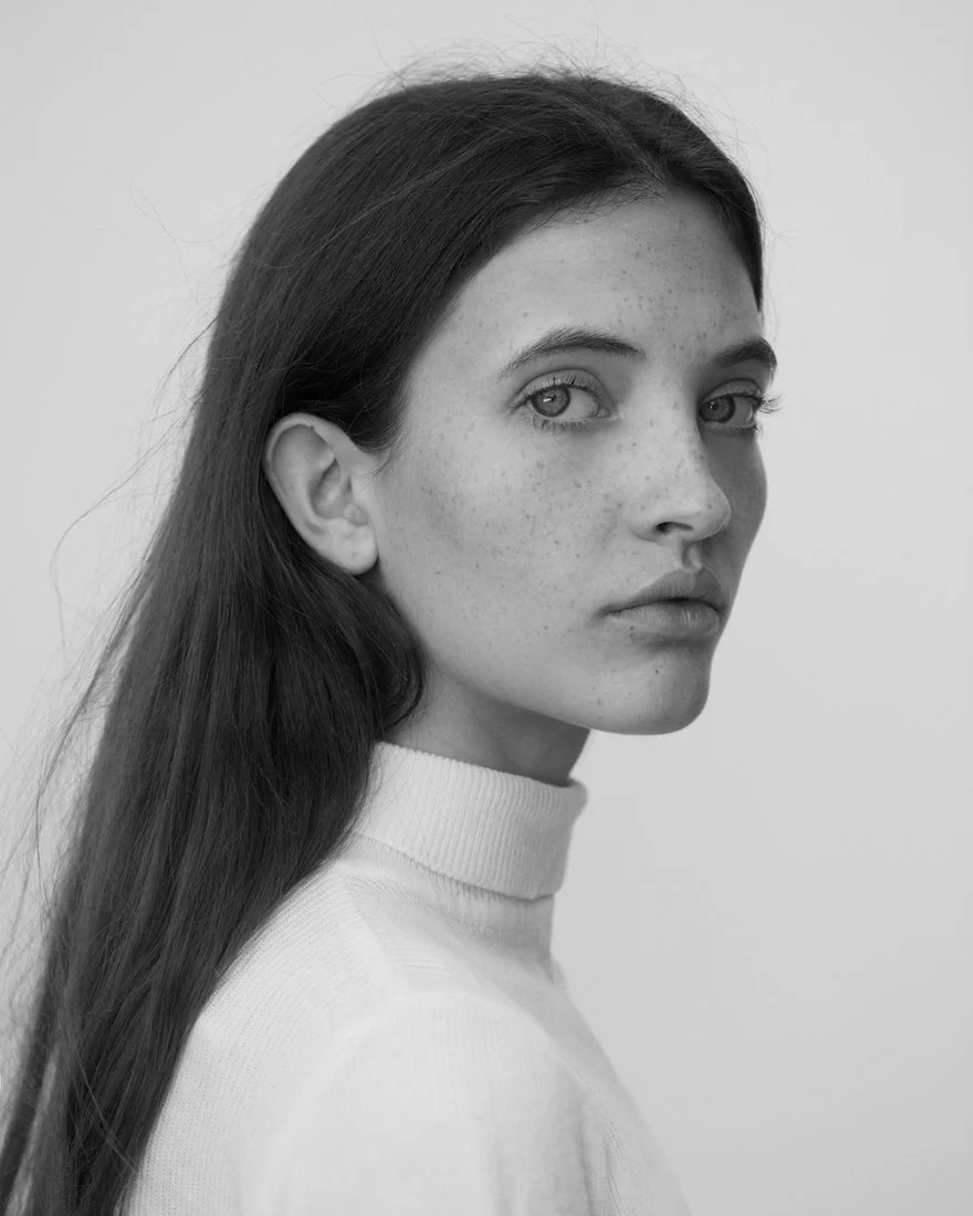 Black and white portrait of a young woman with long dark hair, wearing a high-neck sweater, looking over her shoulder.