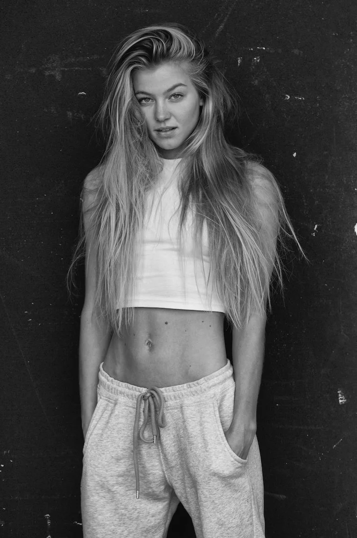 A young woman with long, wavy blonde hair standing against a dark textured wall. She wears a white crop top and light gray sweatpants with a drawstring, with her hands in her pockets.
