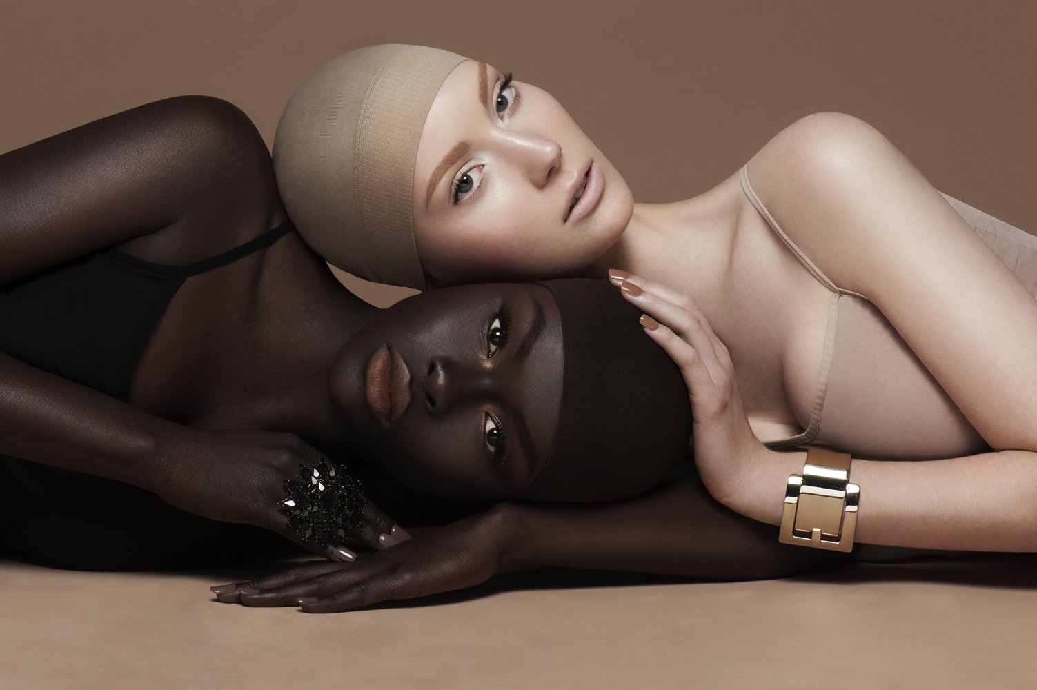 Two women, one with dark skin and one with light skin, lying down together. The dark-skinned woman has her hand resting on the other woman’s cheek, and both are gazing towards the camera. They are styled elegantly with makeup and accessories, one wearing a beige swim cap and a black outfit, the other with makeup and a beige camisole. The background is a neutral brown.
