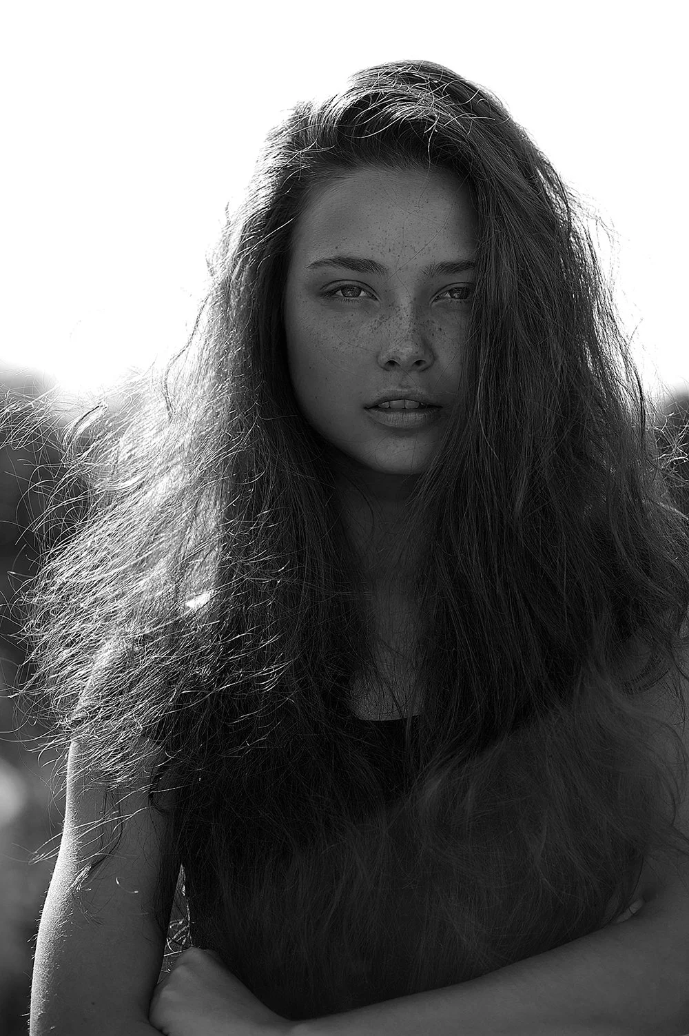 Black and white photograph of a young woman with long, curly hair, looking directly at the camera with a serious expression, outdoors with the sunlight behind her.