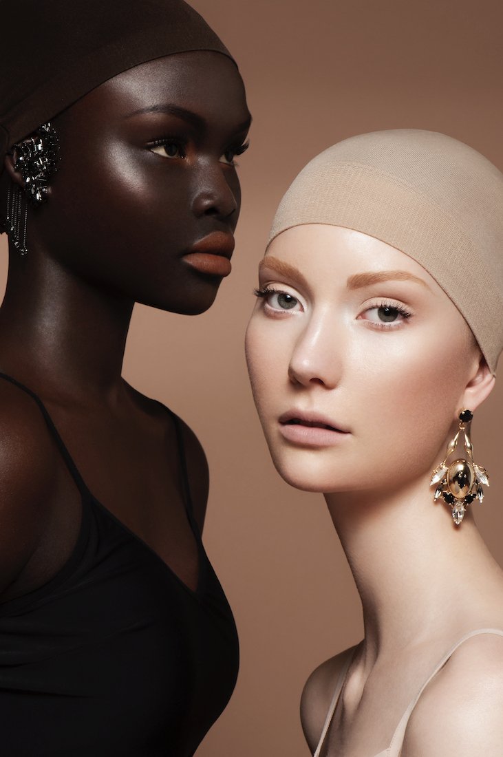Close-up of two women, one with dark skin and the other with light skin, both wearing head coverings and earrings, against a beige background.