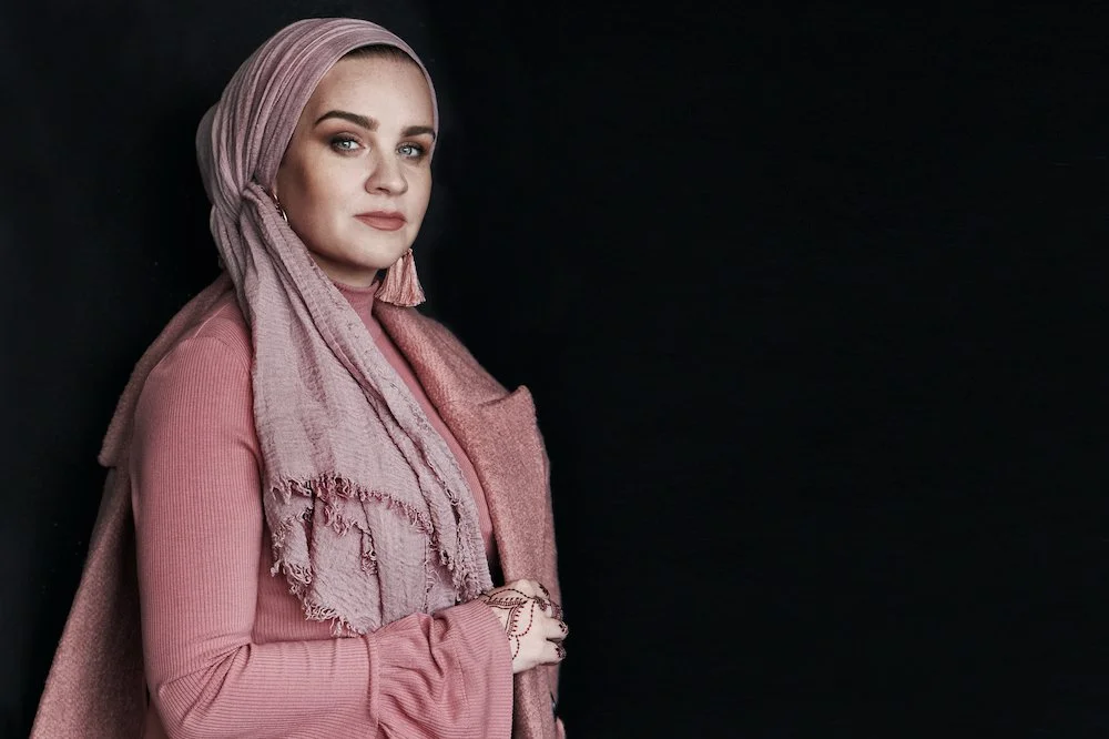 A woman wearing a pink headscarf and matching pink top standing against a black background.