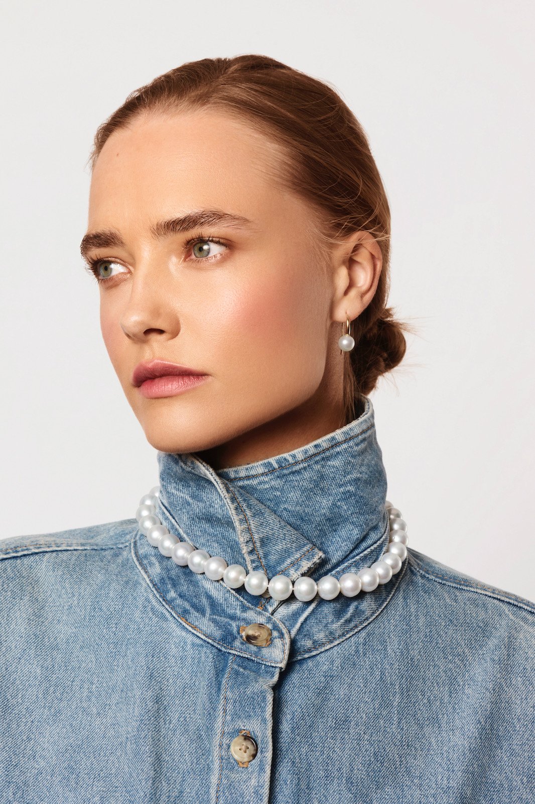 A woman with red hair styled in a low bun, wearing a pearl necklace and pearl earrings, with a denim jacket against a plain white background.