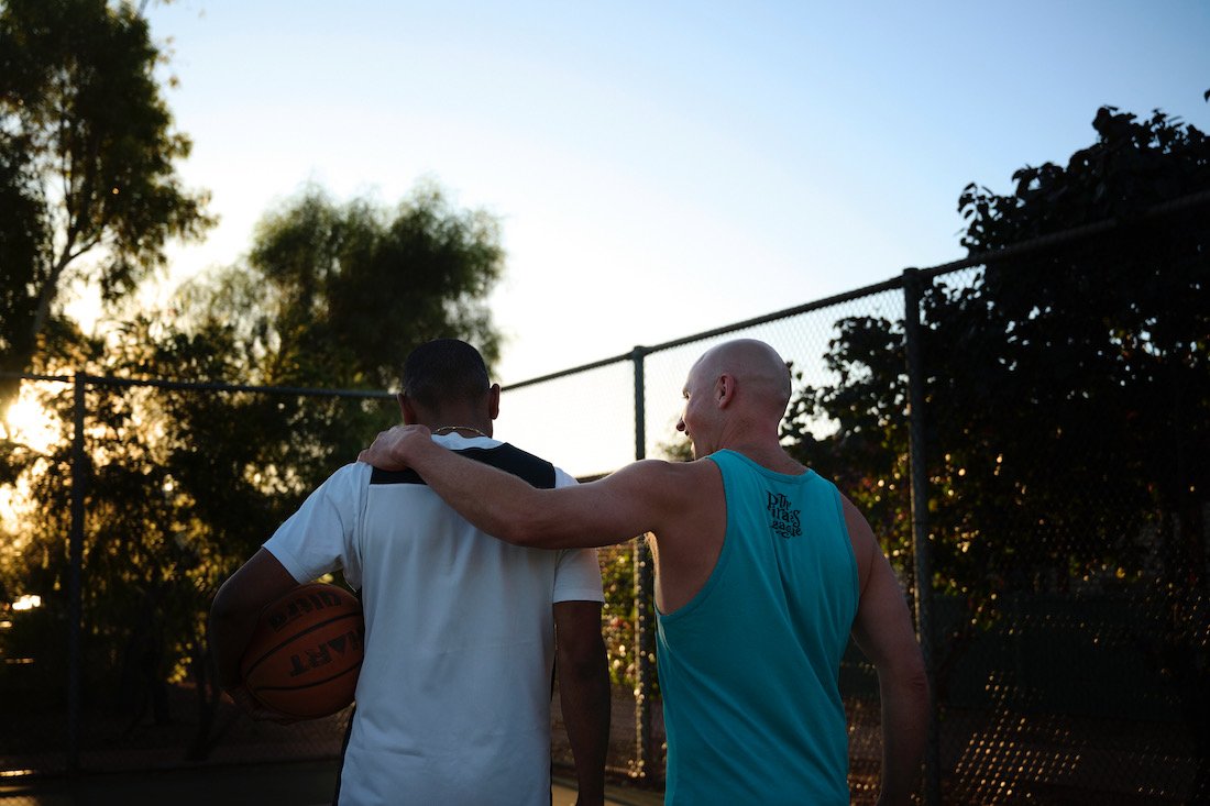 A man with a bald head and wearing a blue sleeveless shirt has his arm around a young man holding a basketball, on a basketball court during sunset.
