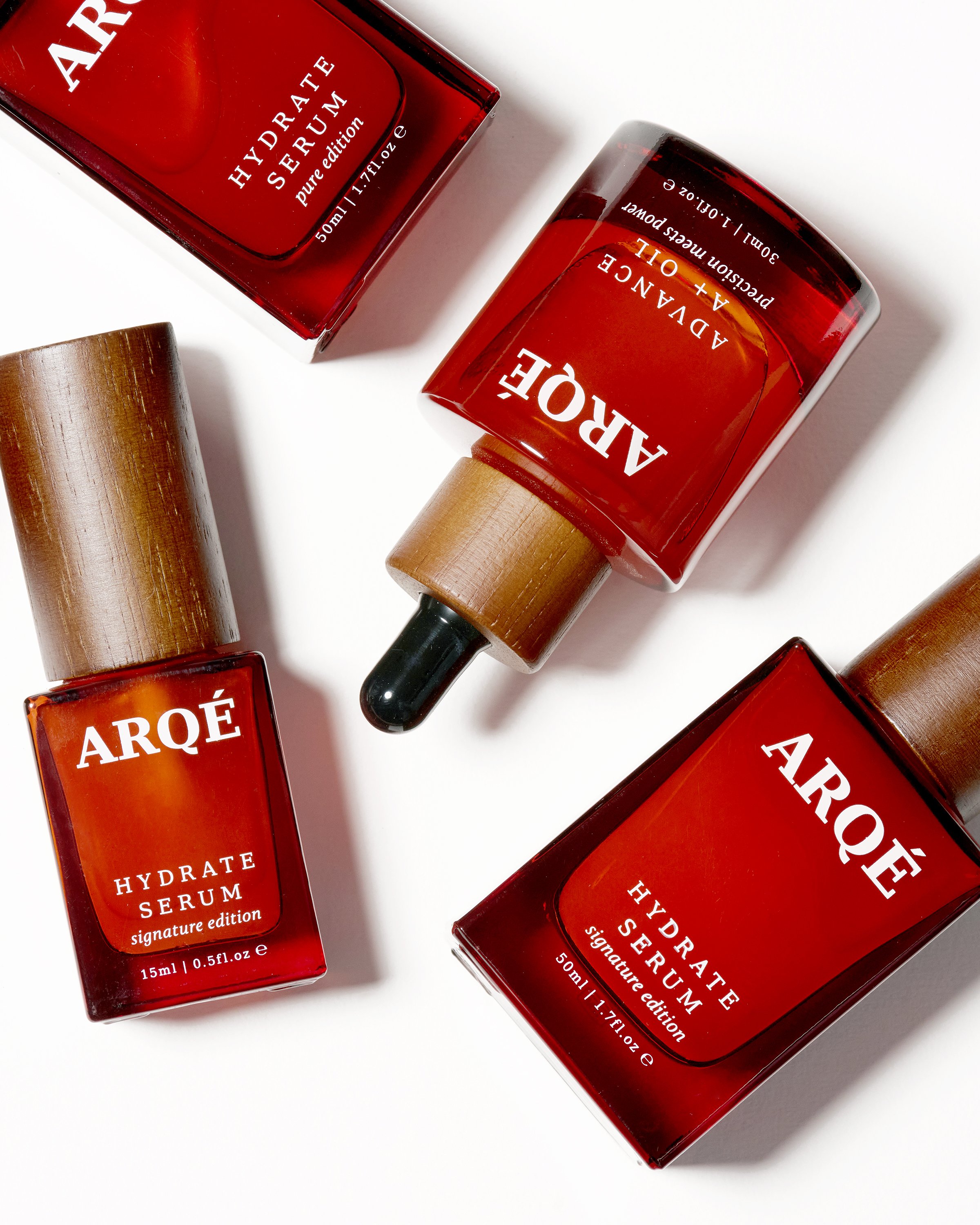 Close-up of three bottles of Arôe Hydrate Serum with red labels and wooden caps on a white background.