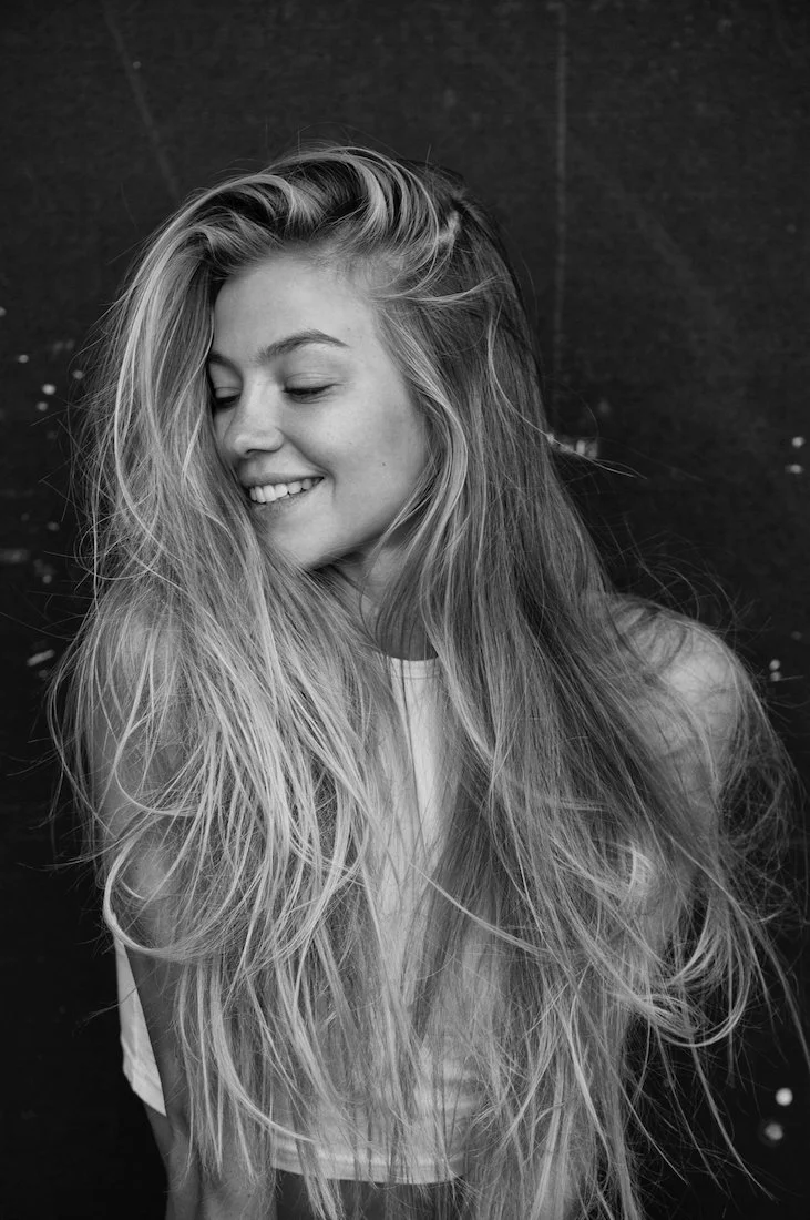 A young woman with long, wavy hair smiling with her eyes closed in front of a dark background.