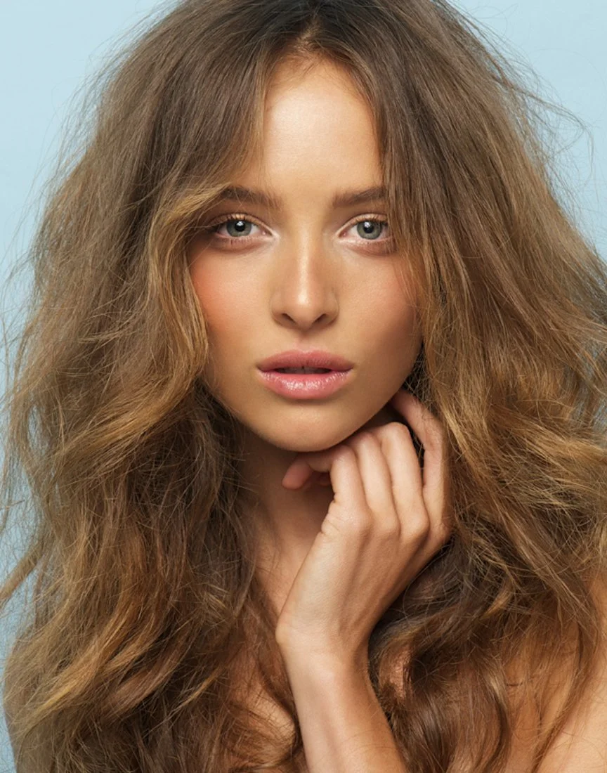A young woman with long, voluminous, wavy red hair, looking directly at the camera with a neutral expression, softly touching her chin with her left hand, against a light blue background.