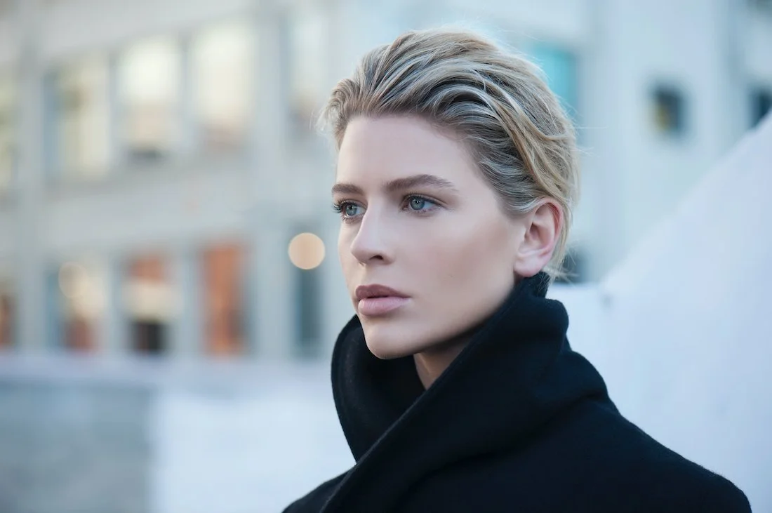 A woman with short blonde hair and blue eyes is wearing a black coat outdoors in an urban area.