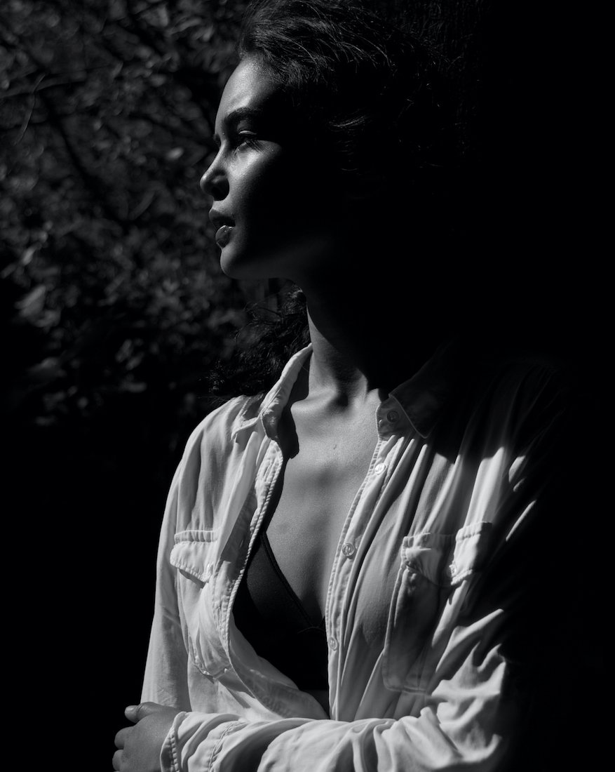 A black and white portrait of a woman in profile, wearing a light-colored shirt with an open collar, looking to the left with sunlight casting shadows on her face and clothing.
