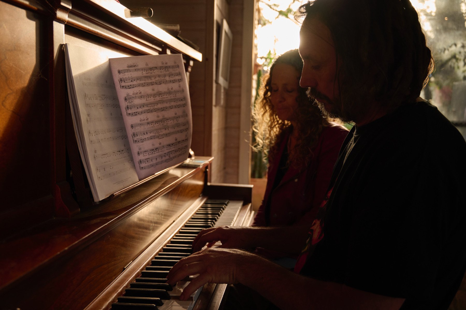 People playing piano together during sunset, with sheet music on the piano, indoors near a window with sunlight streaming in.