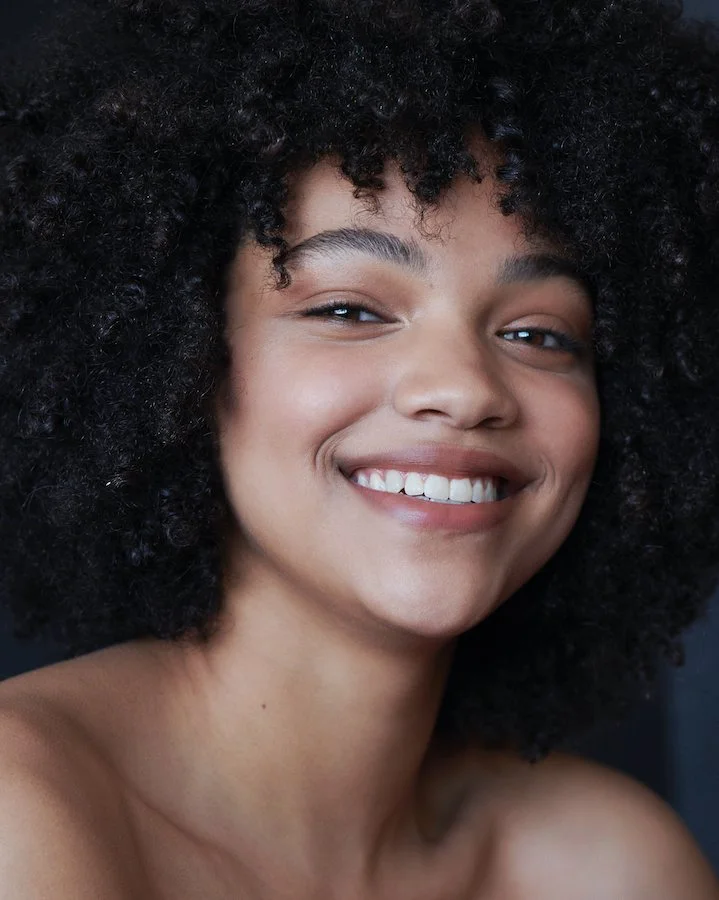 Close-up of a smiling woman with dark, curly hair and minimal makeup, looking directly at the camera.