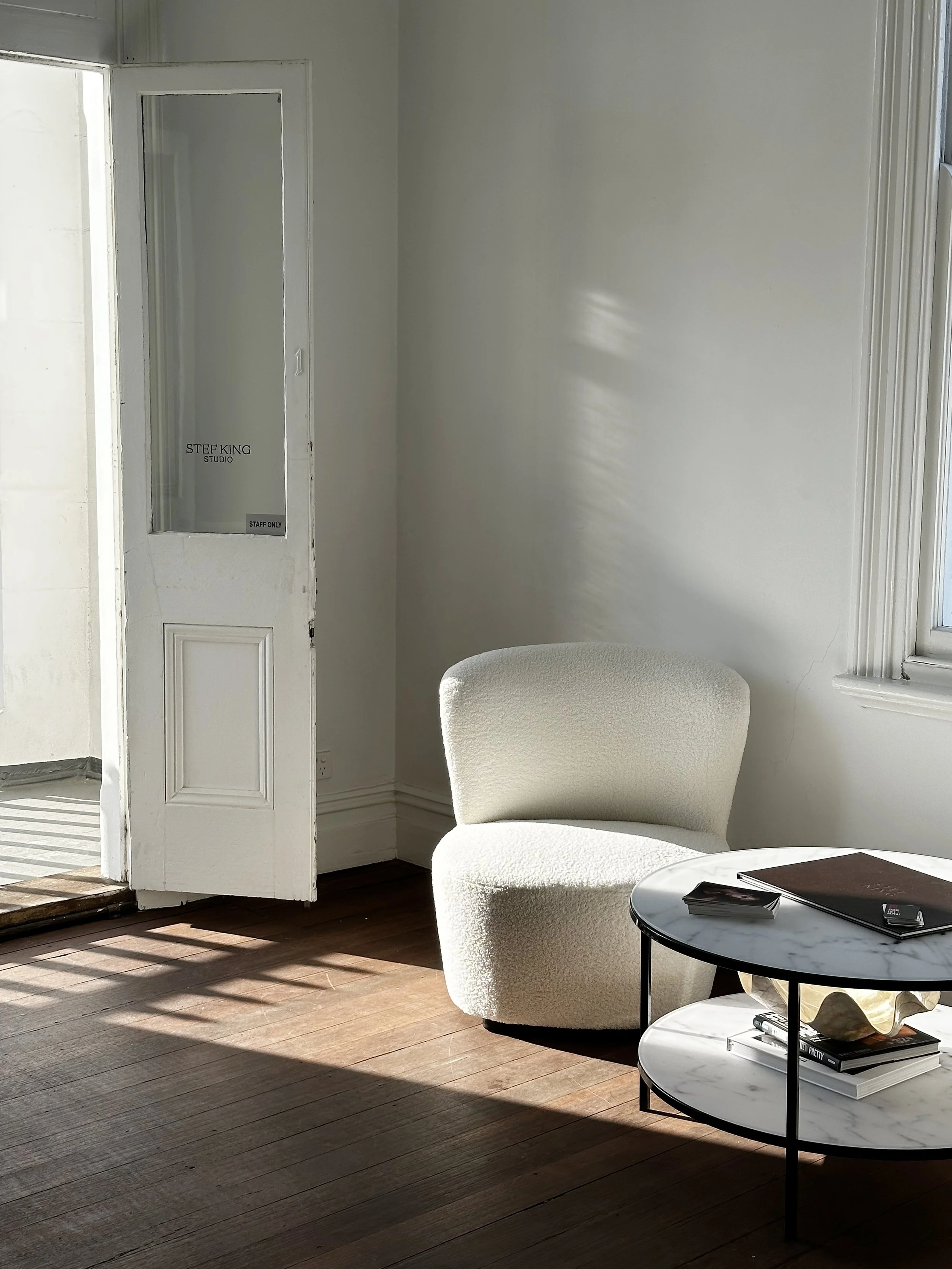 A sunlit room with white walls and a wooden floor, featuring a white upholstered chair and a round marble-top coffee table with books and accessories, an open door labeled 'STEF KING STUDIO' leading to an outdoor space.