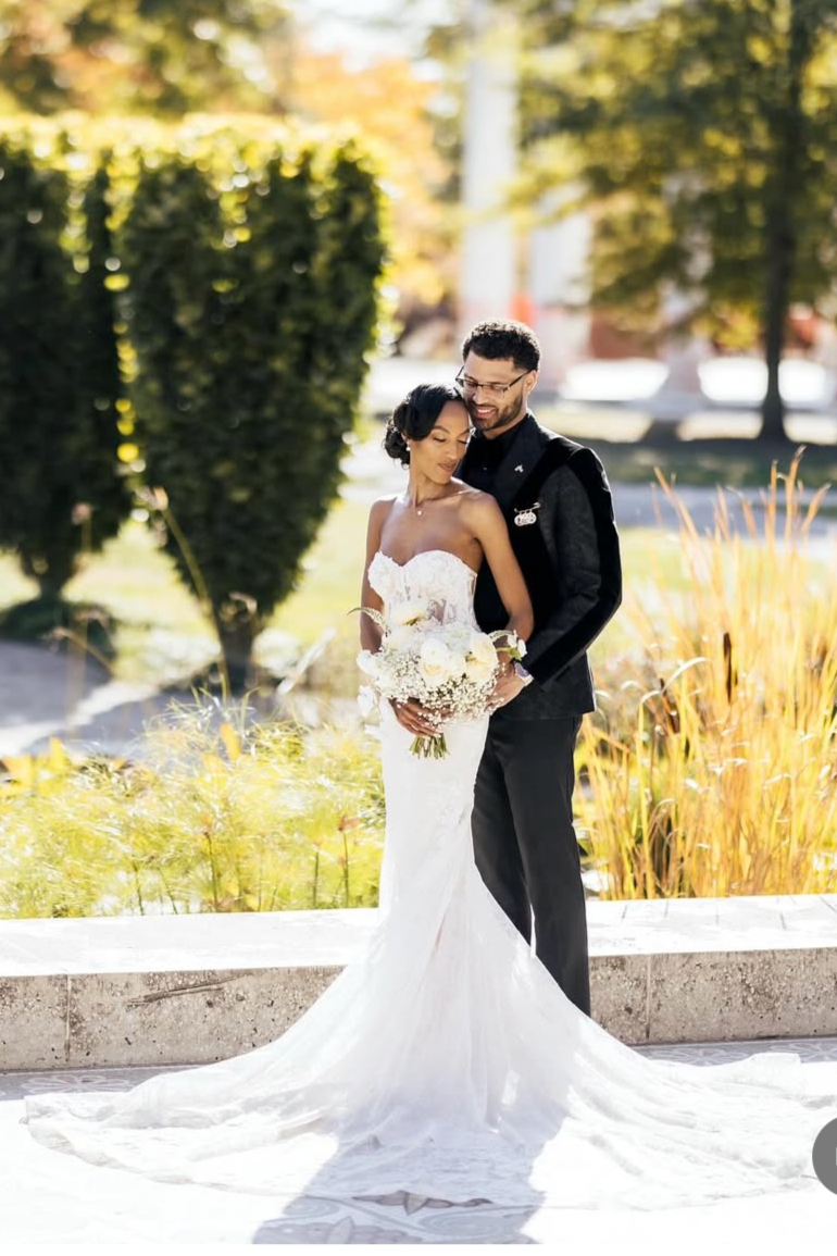 A newlywed couple standing outdoors in a park during daytime. The bride is wearing a white strapless wedding gown and holding a bouquet of white and cream flowers. The groom is dressed in a black tuxedo with glasses. They are embracing each other, smiling, with yellow and green autumn foliage in the background.
