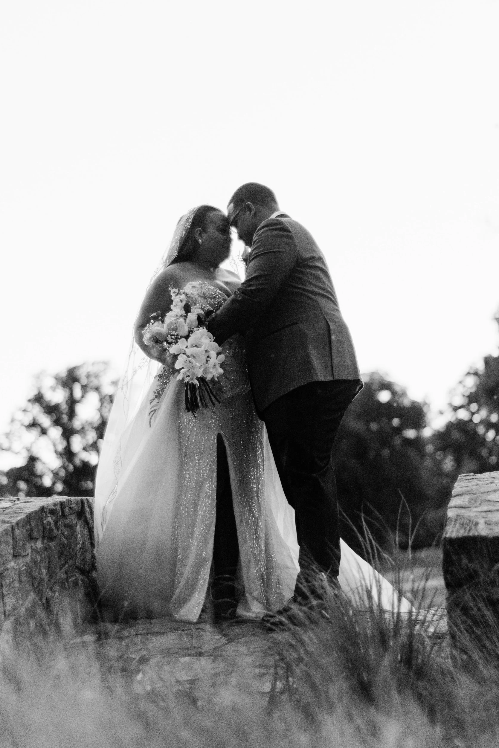 A black and white photo of a bride and groom standing closely, touching foreheads, outdoors on a stone ledge, with trees in the background.