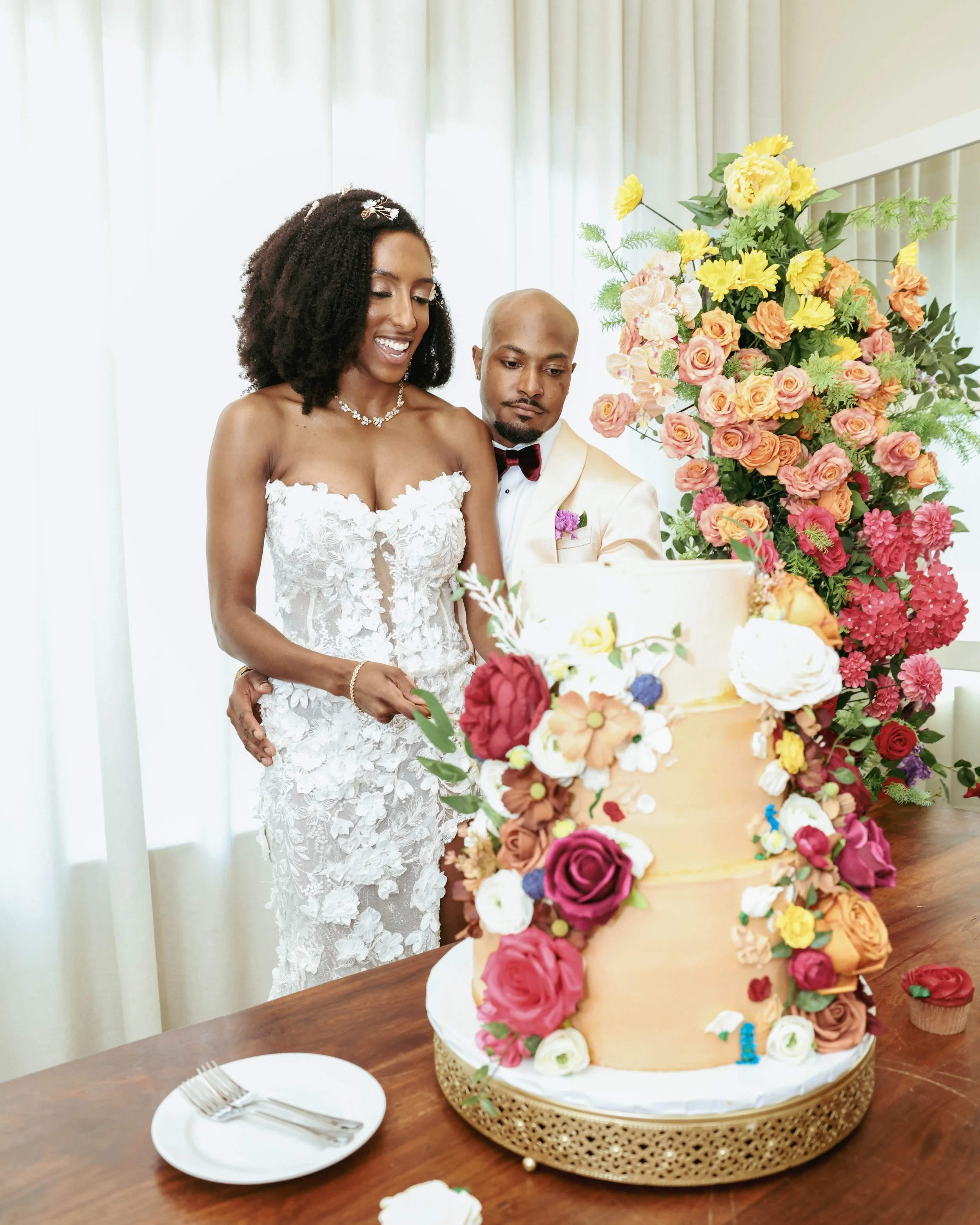 Bride and groom cutting a tiered wedding cake decorated with fresh flowers, with a large floral arrangement in the background.