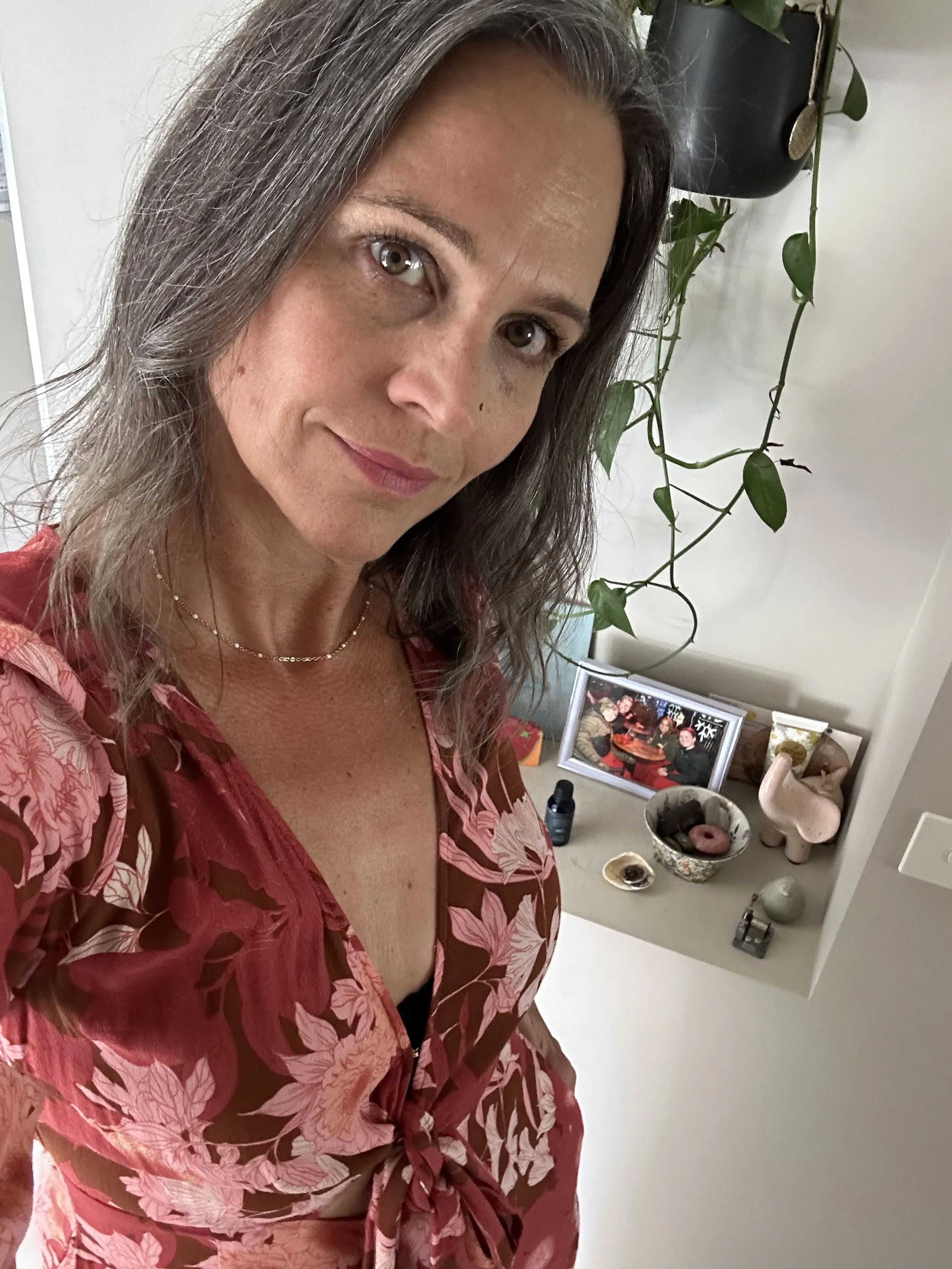A woman with wavy brown hair and light skin looking at the camera, wearing a pink and brown floral dress. Behind her on a white surface are a picture frame, some skincare products, a small bowl with a donut, and various decorative items. A hanging plant is partially visible in the background.