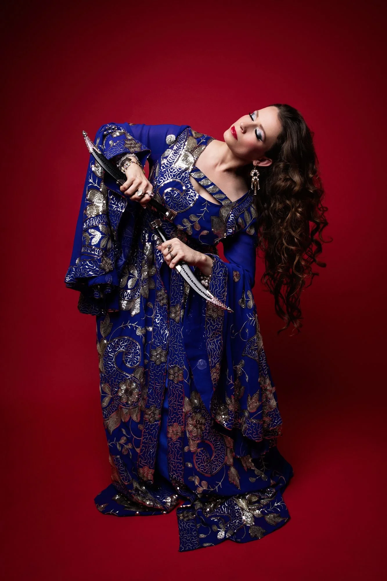 Lara Victoria bellydancer is posed for Kawleeya dagger dance. She wears a lavish blue and gold dress and holds two daggers.