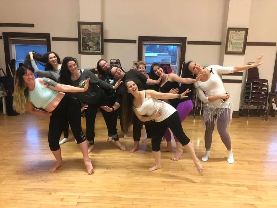 A group of women, led by Olympia Belly Dancer Lara Victoria, are leaning sideways in position for Kawleeya Iraqi dance.  They are in a belly dance studio in Lacey. They are smiling and having fun.