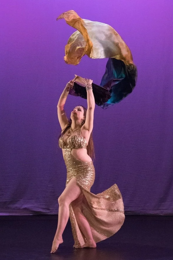 Olympia belly dancer in a glittery gold dress performing at Broadway theater in Seattle Washington, holding colorful fan veil above her head with a purple background.
