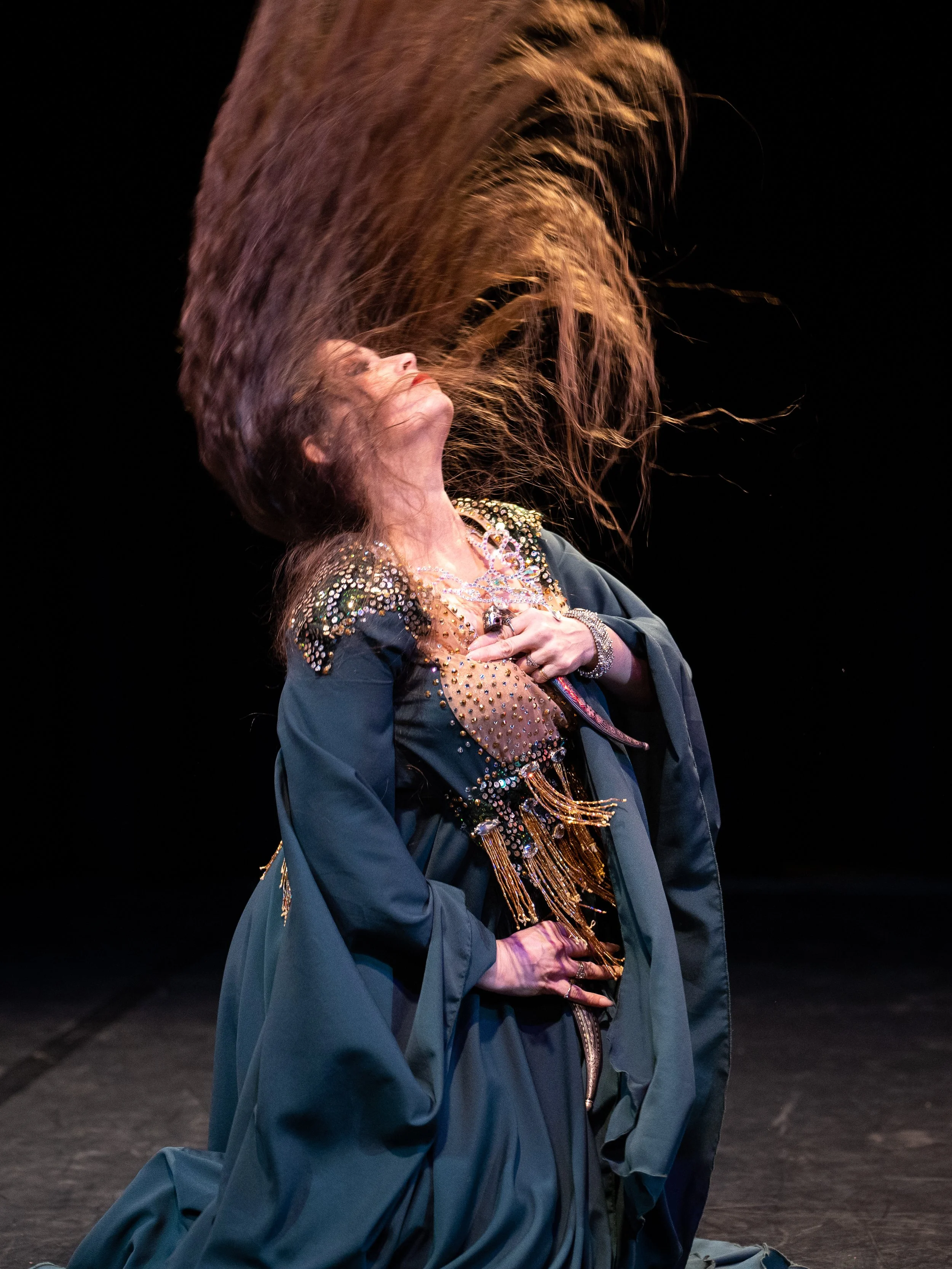 Lara Victoria bellydance performs Iraqi Hacha'a. She kneels with daggers in a green dress. She flings her hair back. The image captures her hair flying through the air.