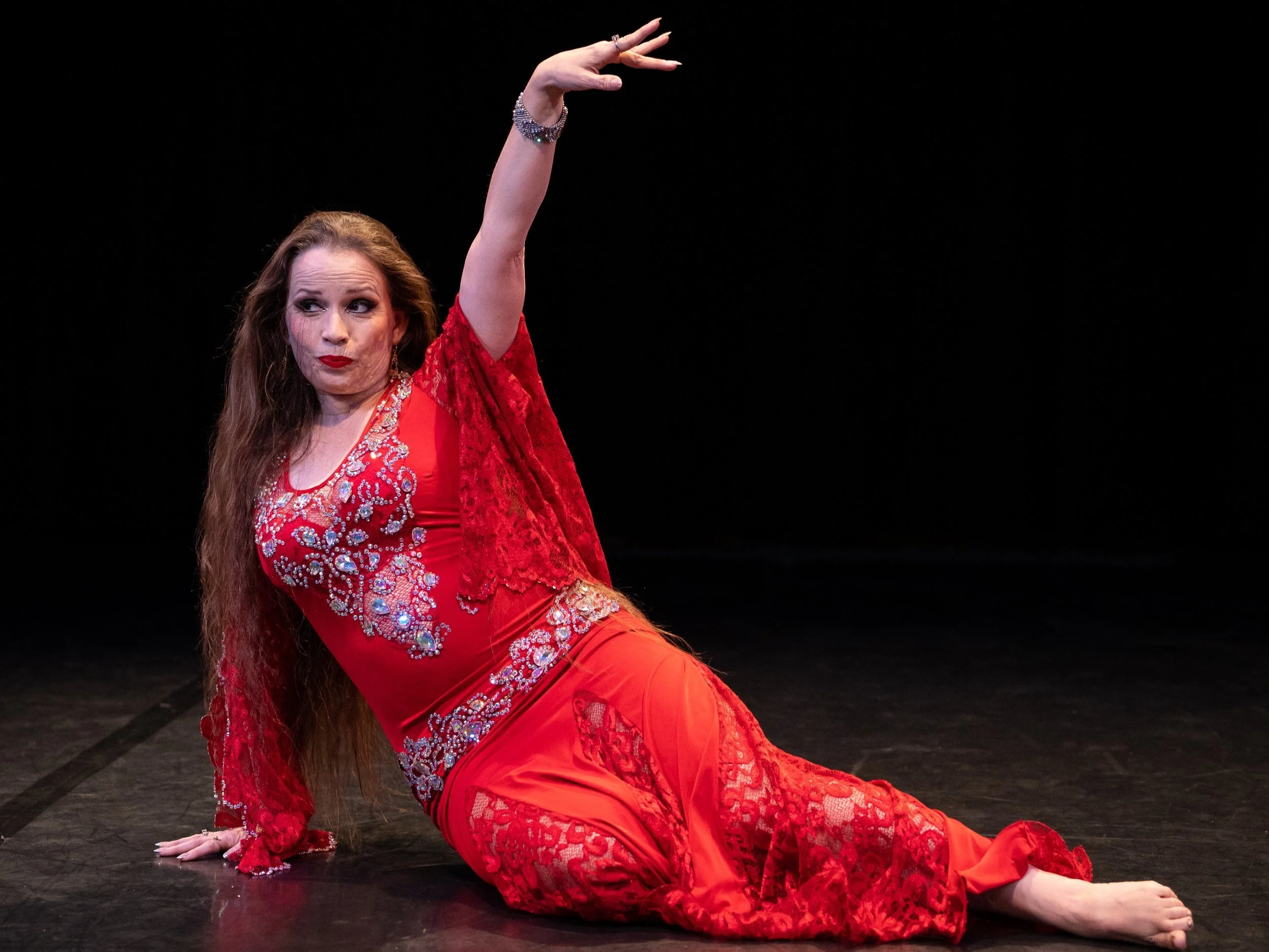 Lara Victoria bellydance performs Kawliya dance in red dress. She is in the floorwork part of her performance. She reclines on the floor and poses with an arm up.