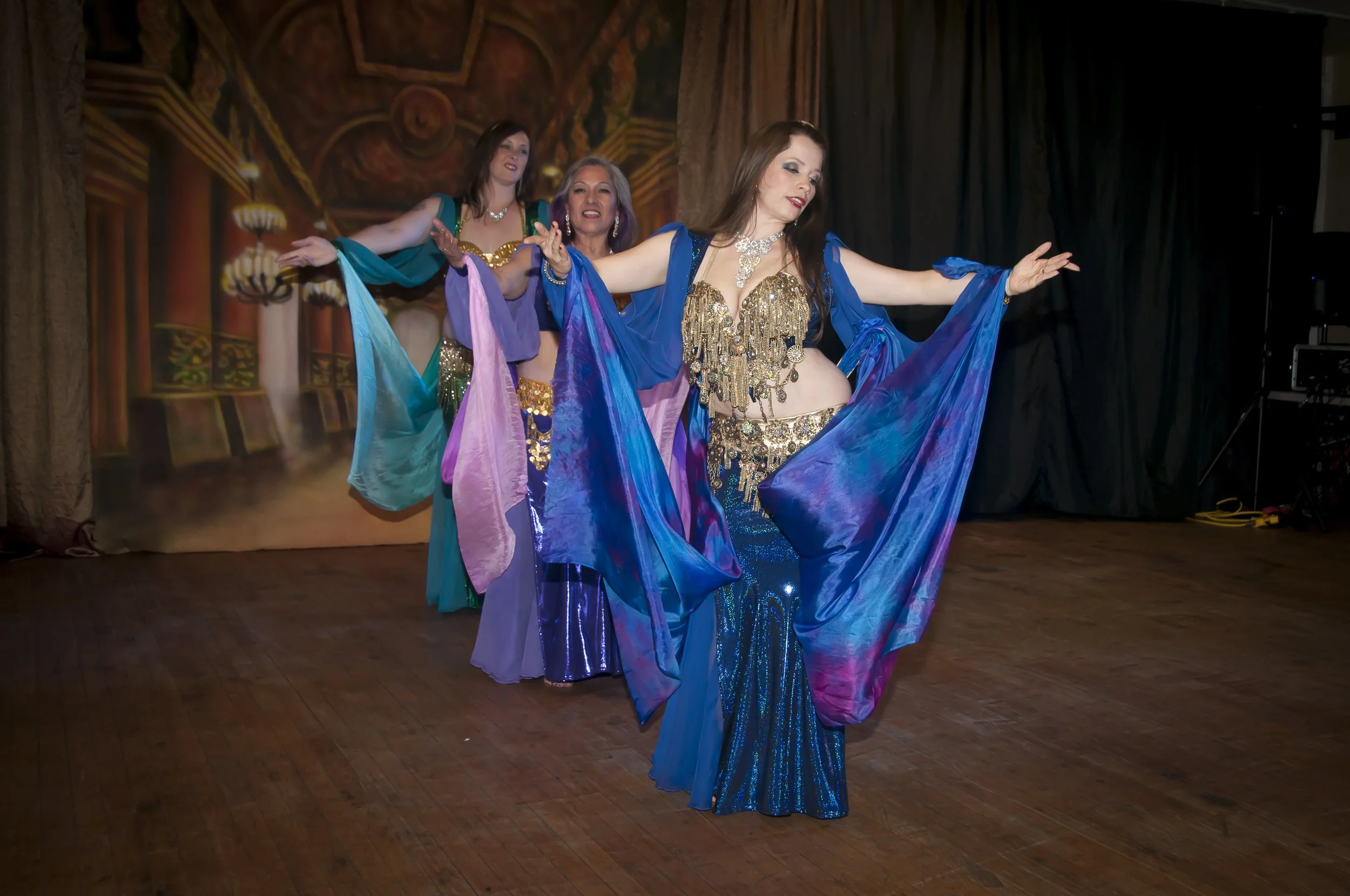 Three belly dancers perform at a bellydance event. They are wearing blue, green & purple skirts with gold coin bedlahs. Lara Victoria is the first bellydancer in the lineup. She teaches in Olympia Washington.