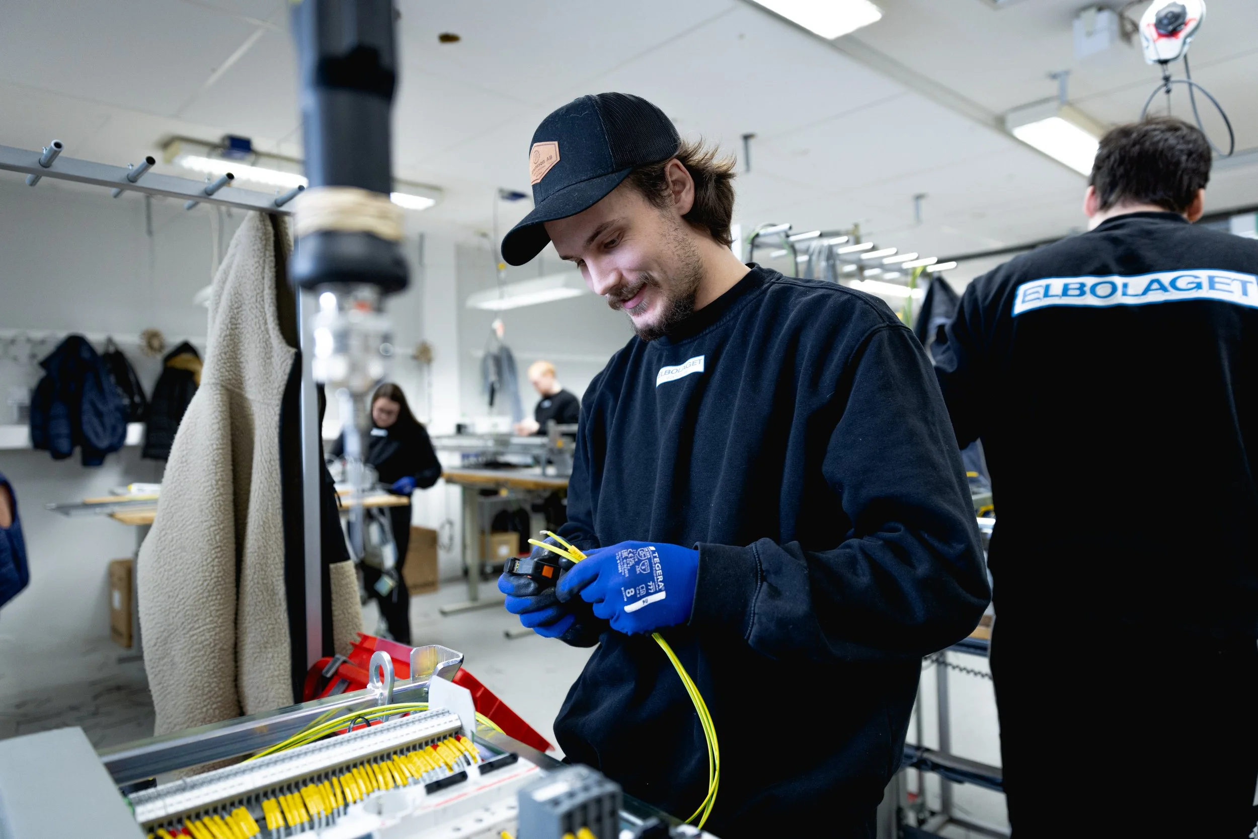 En närbild på en man med skägg och svart keps som ler medan han arbetar med gula kablar i en verkstadsmiljö. Han bär blå arbetshandskar av märket Tegera och en svart tröja.
