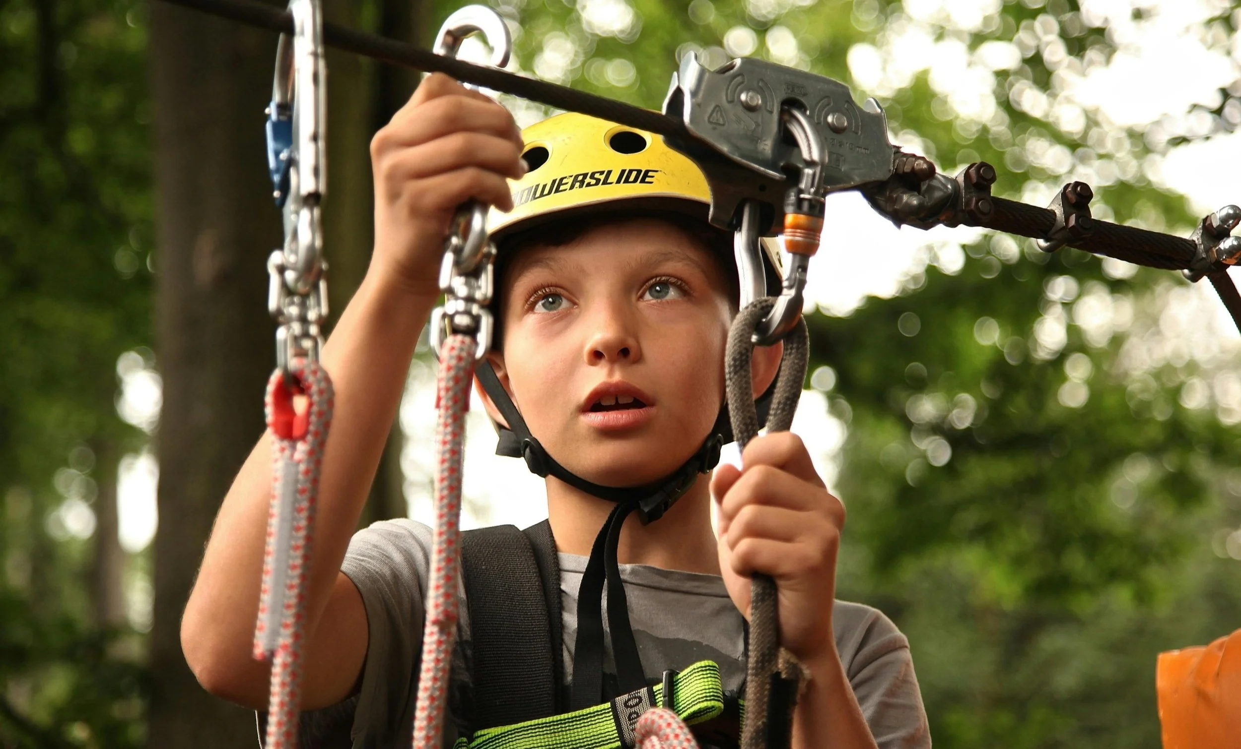 A young boy attaches a climbing rope to another rope with a metal clip. He wears a yellow helmet and a look of concentration on his face. Trees fill the background.
