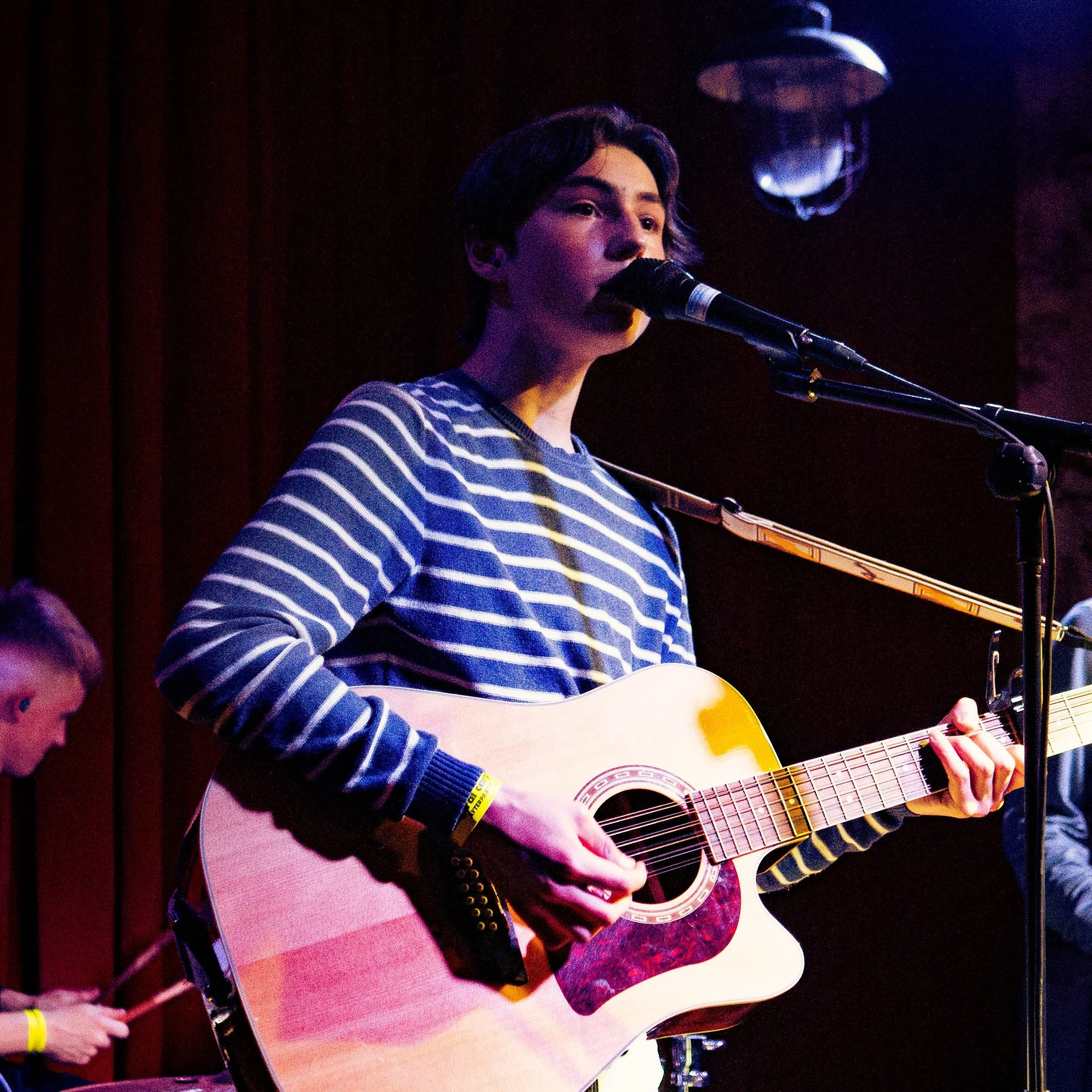 John Denton stands on stage. He strums his acoustic guitar while singing into a microphone and looking out to the crowd.