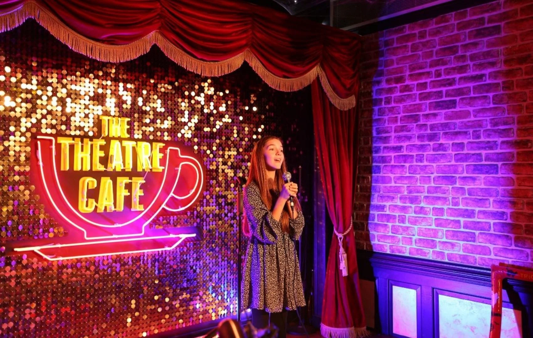 Young teen Eva Johnson sings into a mic on a stage. A neon sign behind her says: The Theatre Cafe. Red stage curtains and shimmering sequins make up her backdrop.