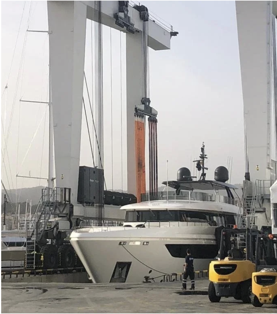 A superyacht suspended beneath a travel lift in a shipyard setting, surrounded by cranes and ground crew.