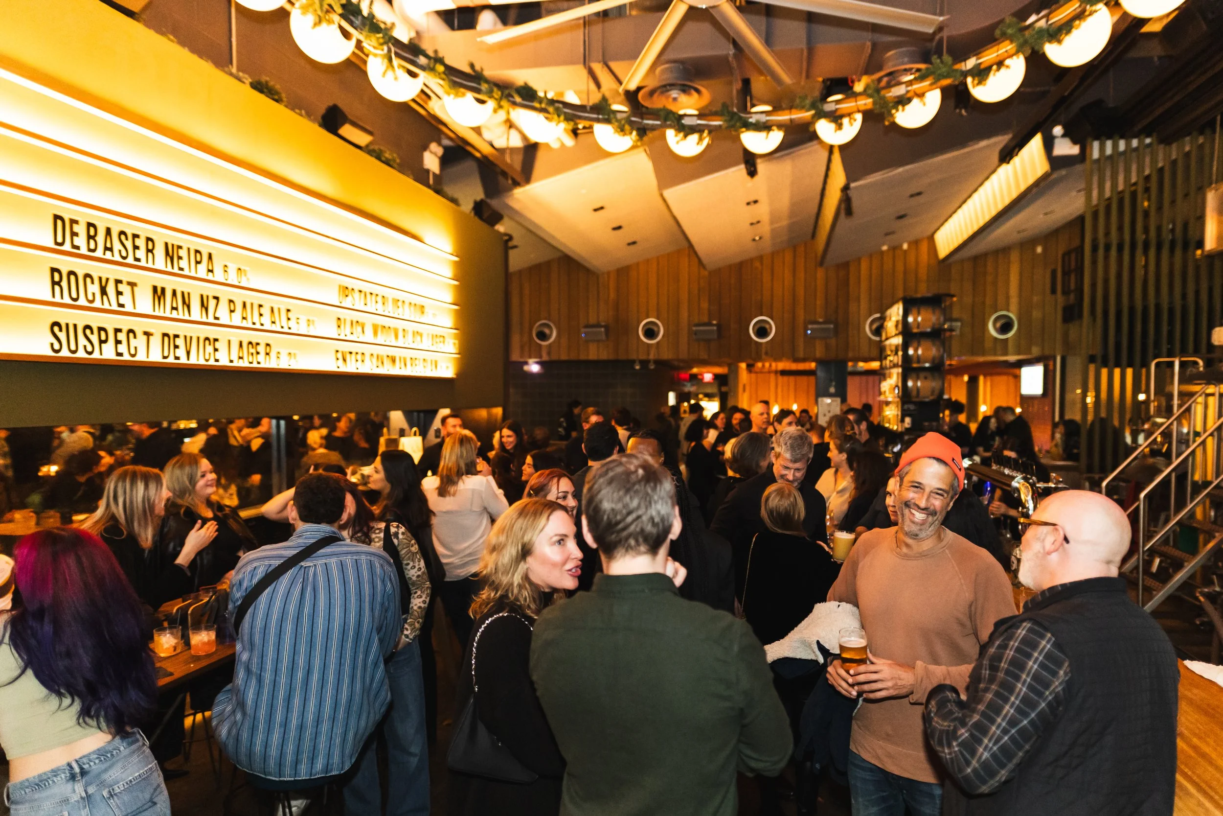 A crowded bar scene with people socializing and a marquee displaying beer names.