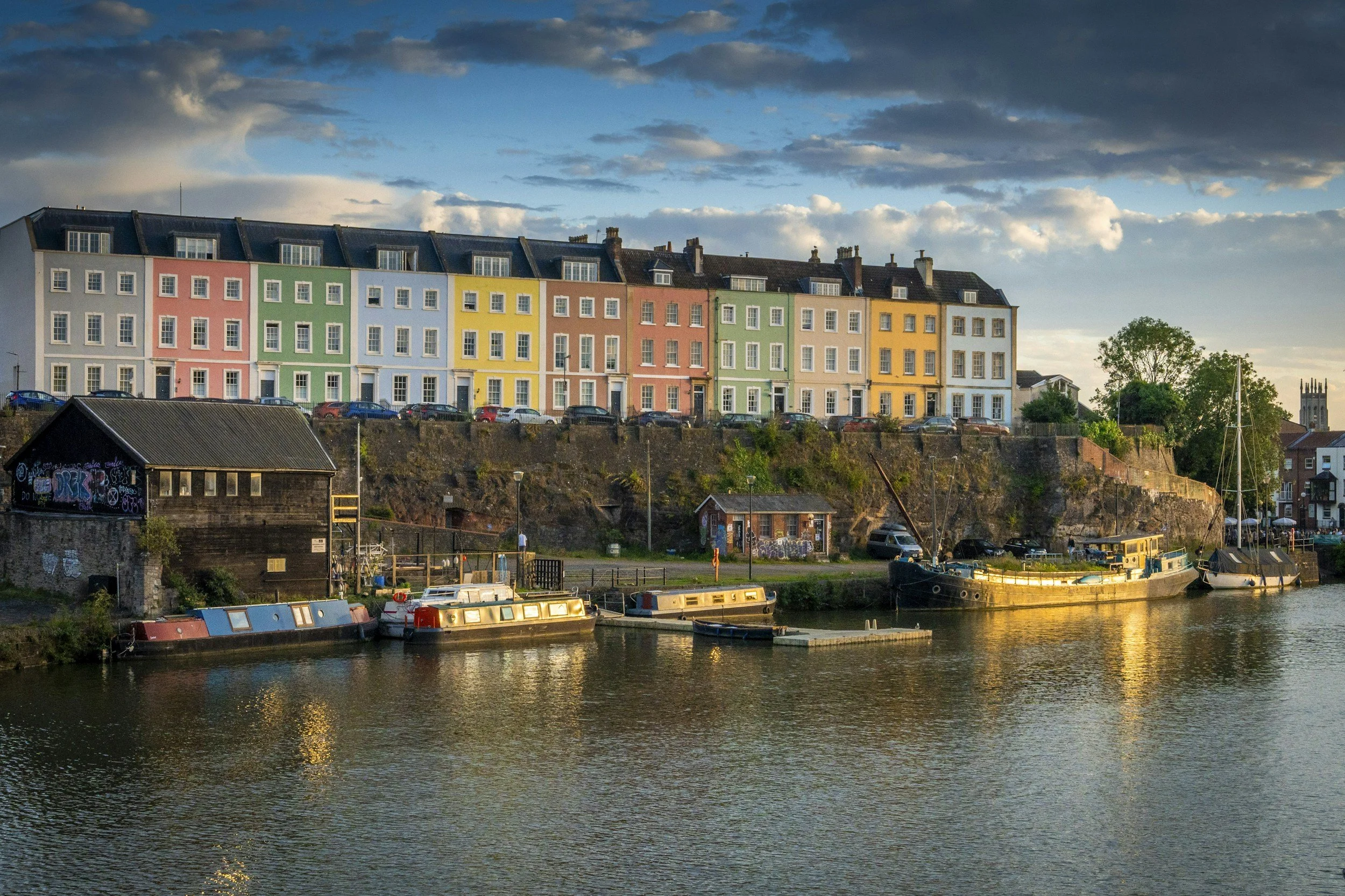 Colorful row of houses on a hill above a river with boats docked at the shore during sunset.